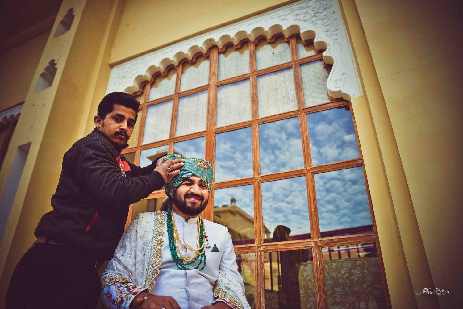 Groom in traditional Indian wedding attire being dressed by assistant near ornate latticed window.