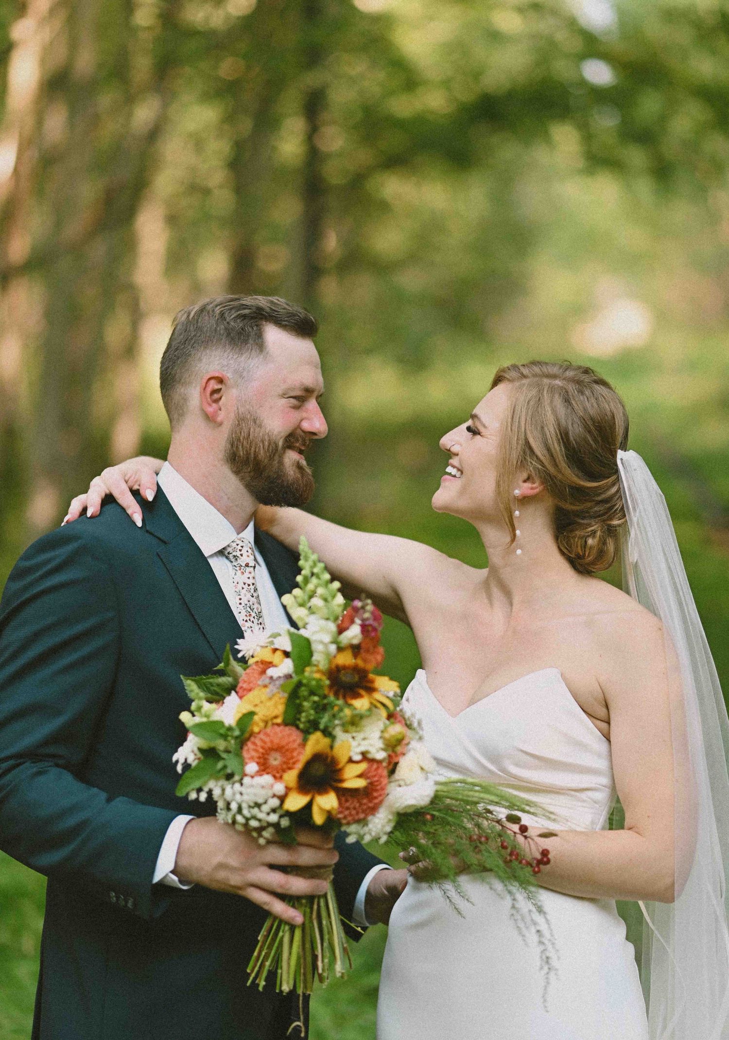 Happy bride and groom smiling together outdoors holding vibrant sunflower wedding bouquet in lush green forest.