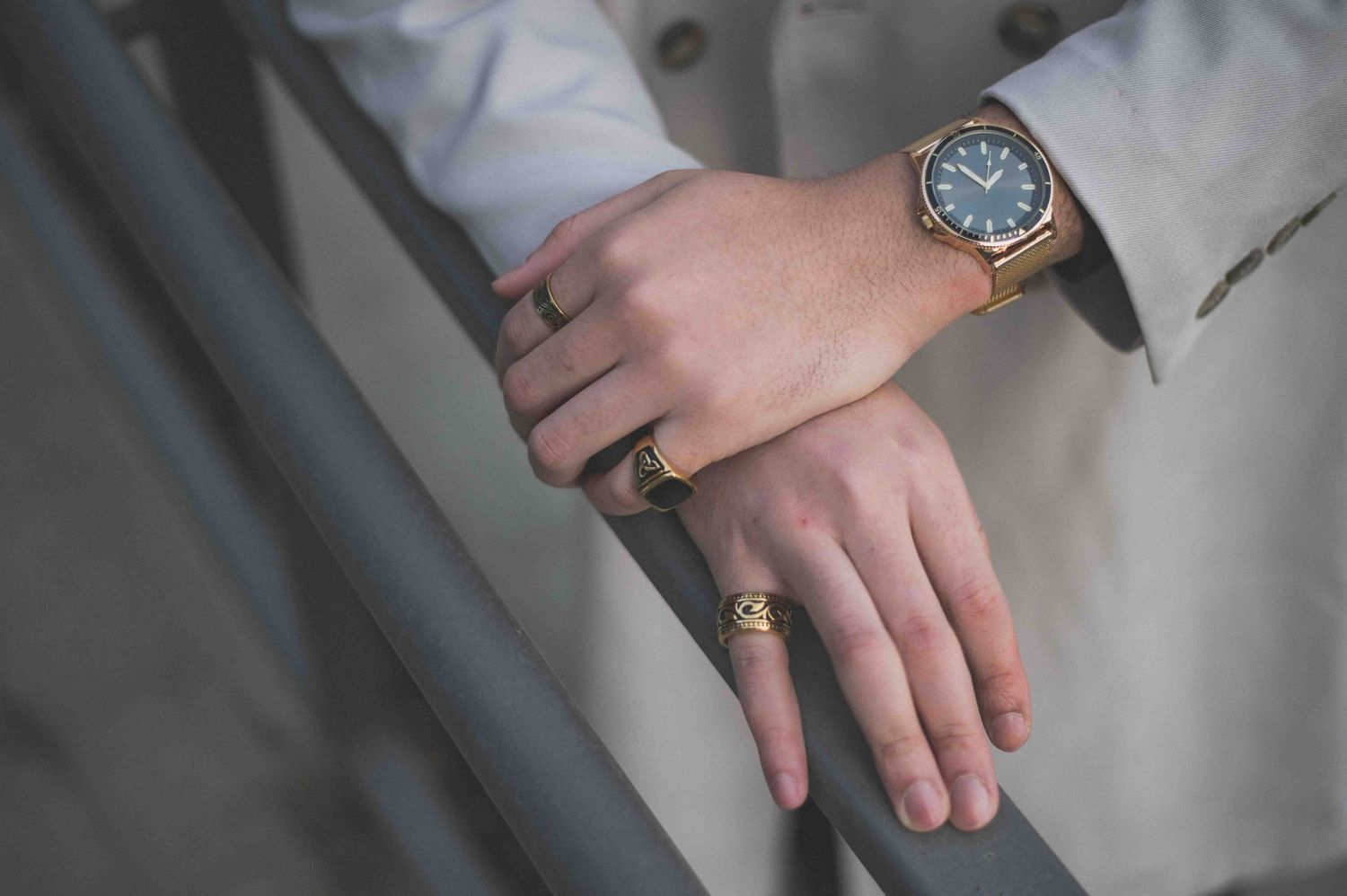 Close-up of hands wearing gold rings and a blue-faced watch with gold bracelet on metal railing.