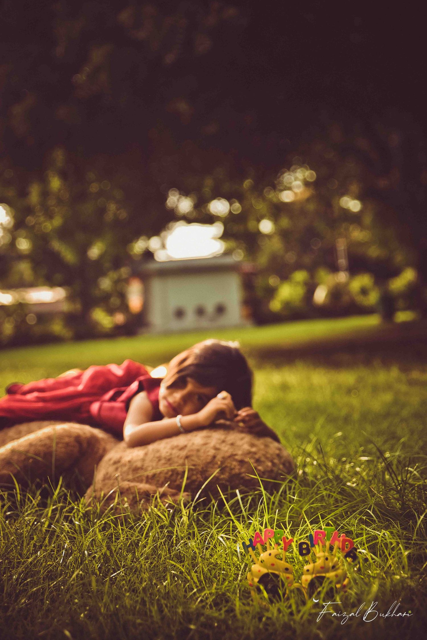 A child in red dress lying on a giant teddy bear on grass with a colorful toy in foreground.