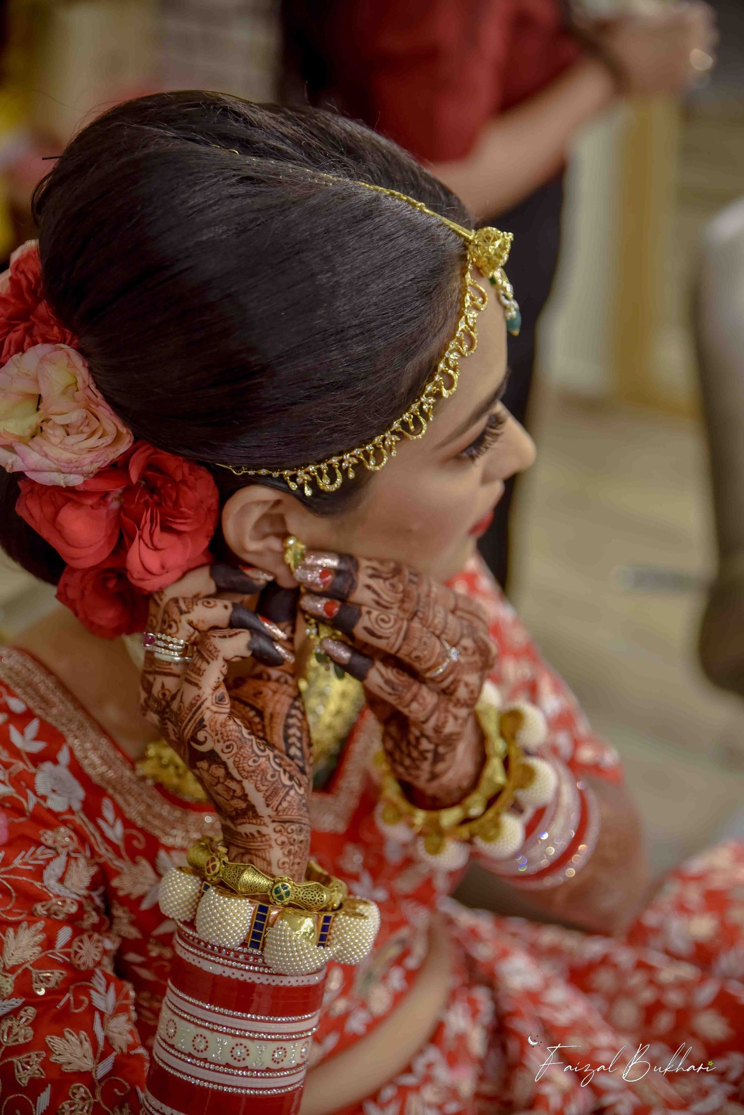 Indian bride in red lehenga adjusting earring with mehndi-covered hands and floral hair accessories.