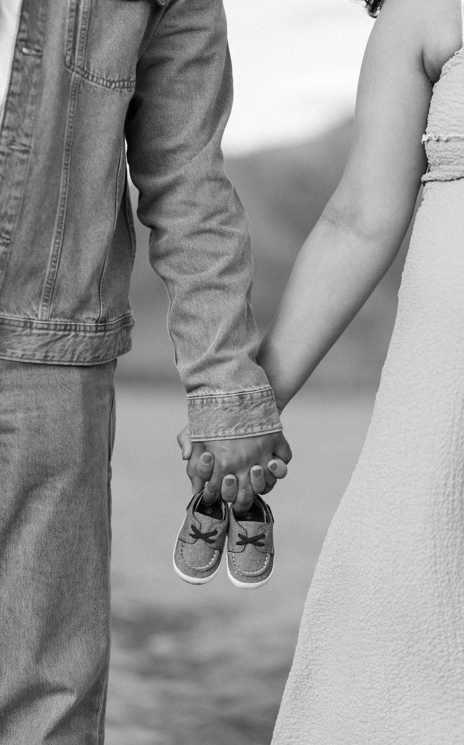 Couple holding tiny baby shoes while holding hands in black and white maternity photo.