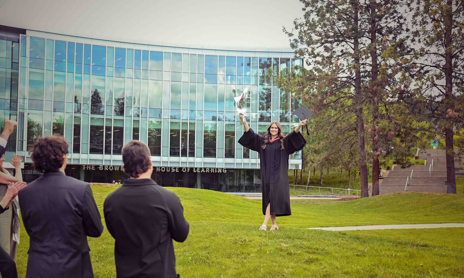 Graduate in cap and gown poses for photos outside modern glass university building on campus.
