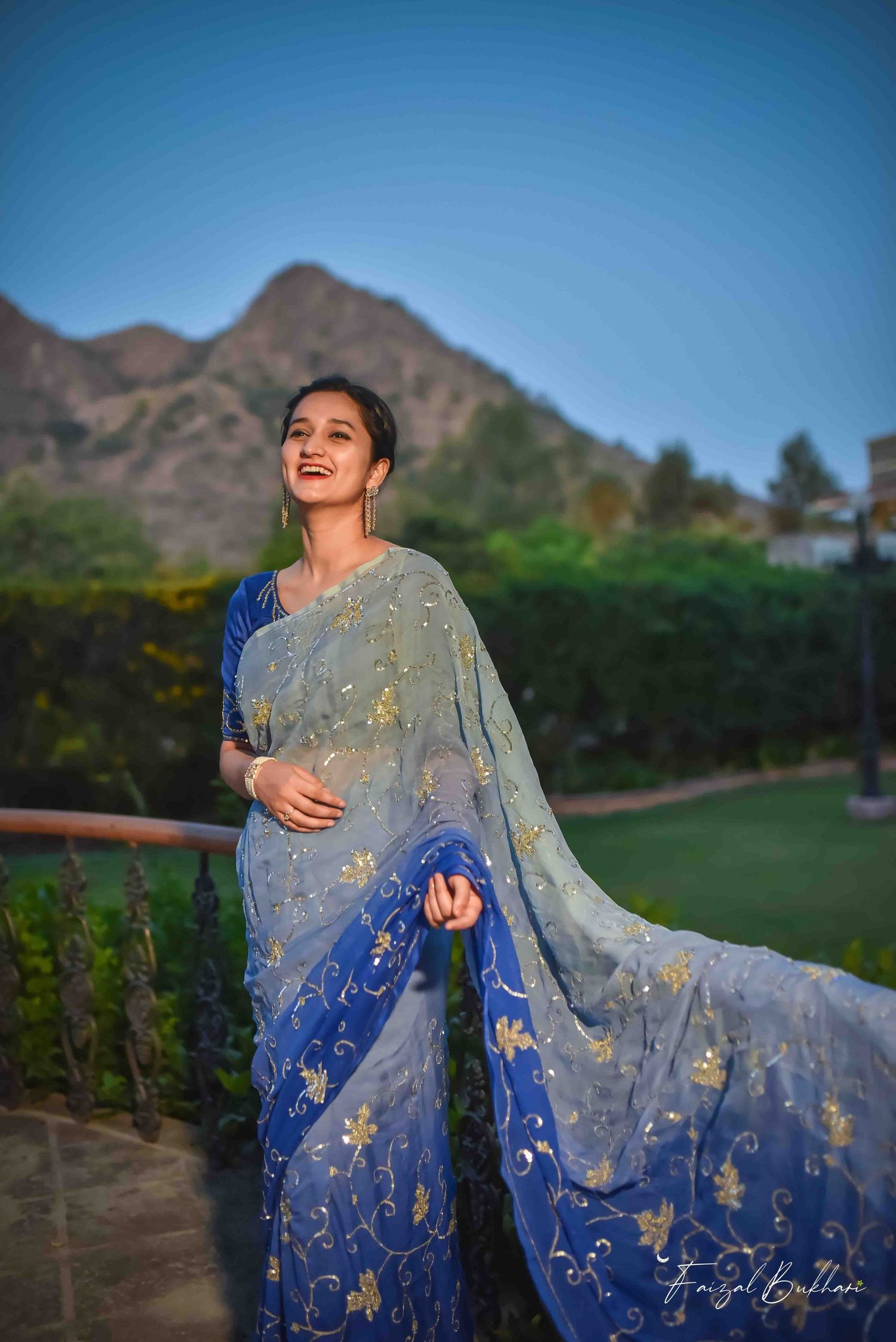 A smiling woman in a blue ombre embroidered saree stands on a balcony with mountains in background.