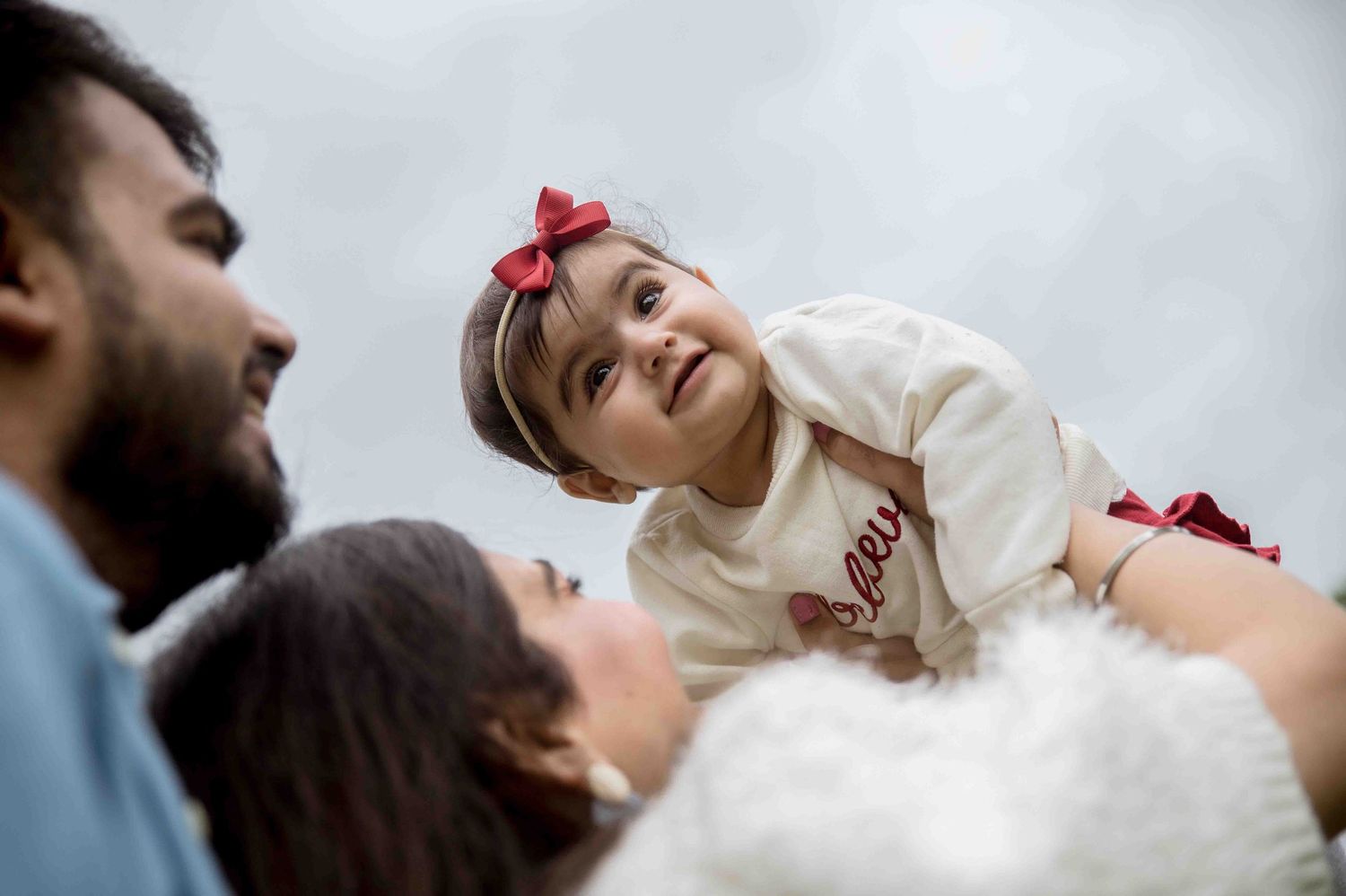 Parents lovingly lifting their smiling baby girl with a red bow in the air.