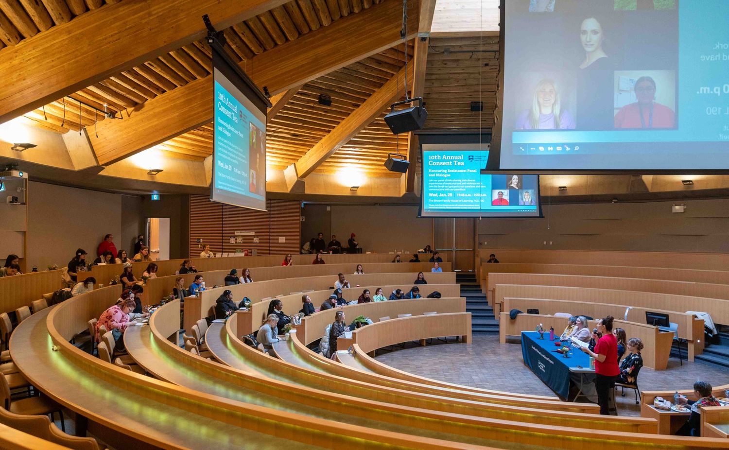 Large auditorium panel discussion with hybrid virtual attendees on screens.