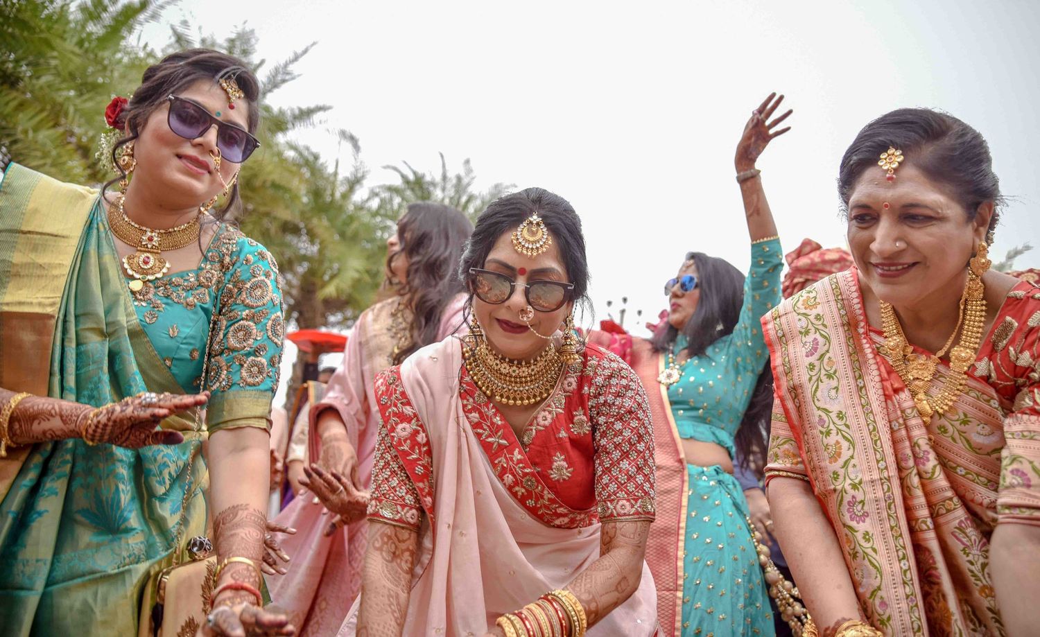 An Indian bride in pink lehenga walking with smiling bridesmaids in colorful sarees at wedding.