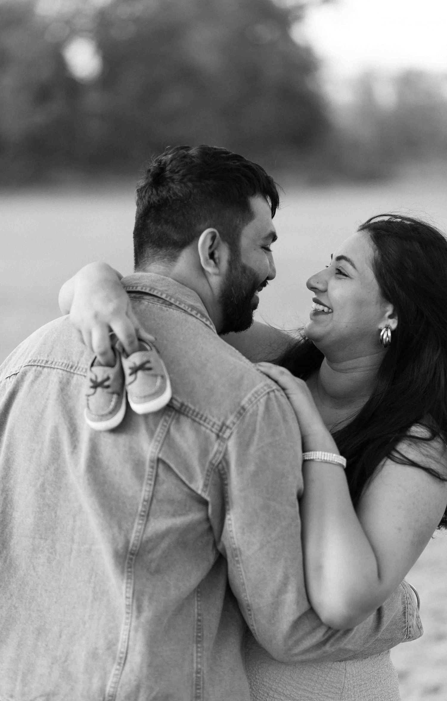 Couple laughing together holding baby shoes in black and white maternity portrait.