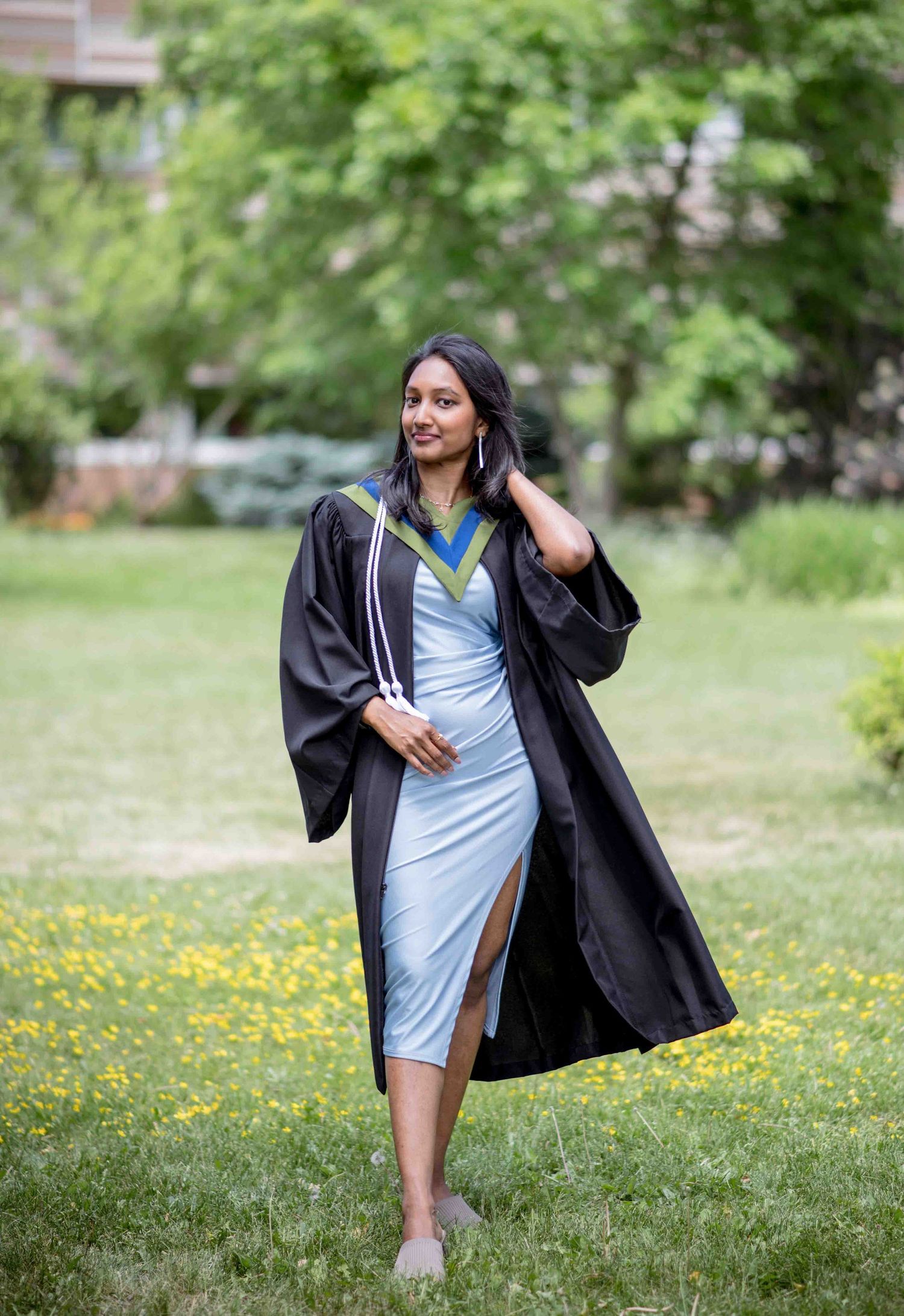Young woman in graduation gown and blue dress walks gracefully across green university campus lawn.