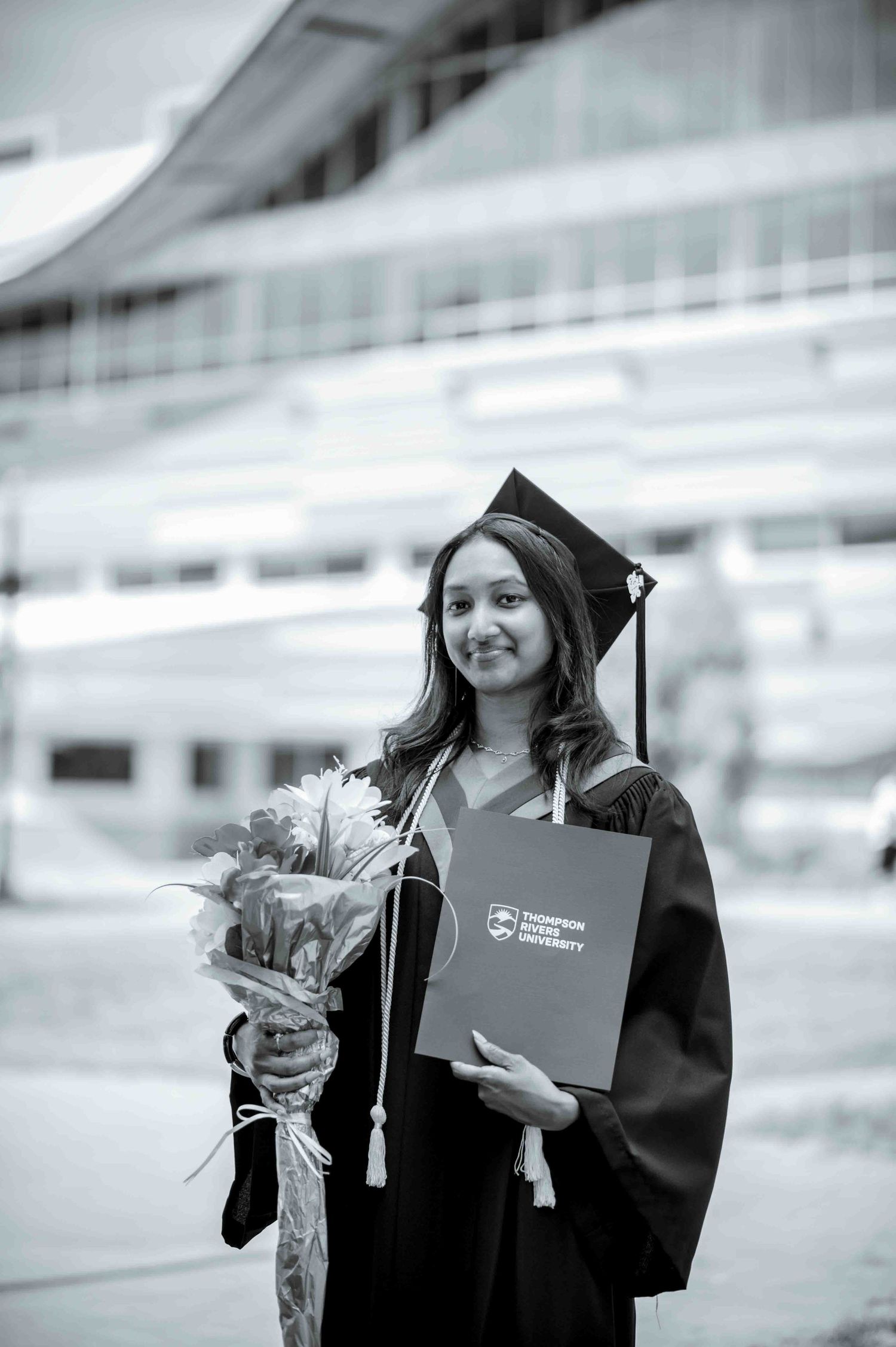 Graduate in cap and gown holds TRU diploma and flowers in classic black and white portrait.