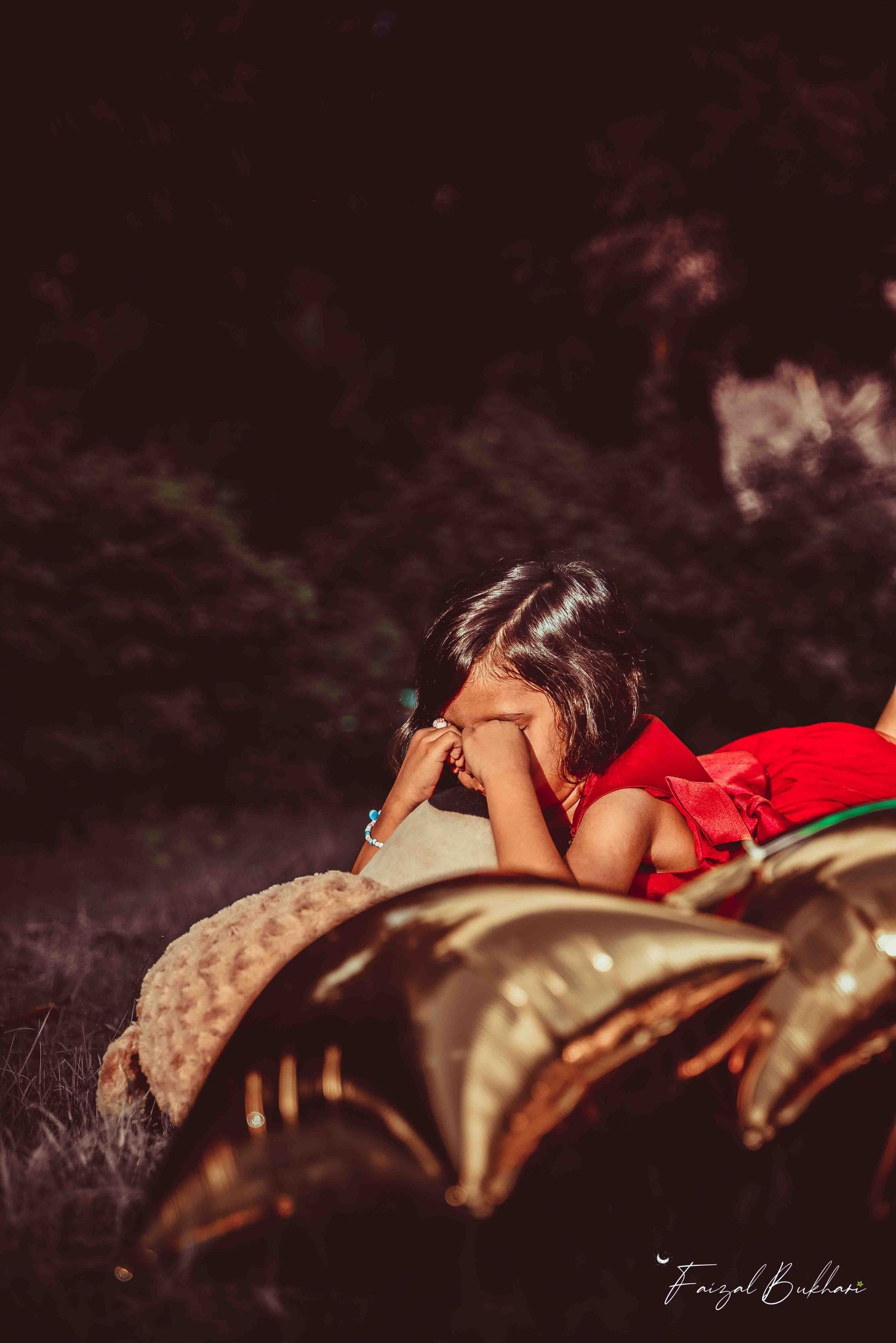 A young girl in a red dress rests on a blanket beside gold star balloons in a moody outdoor setting.