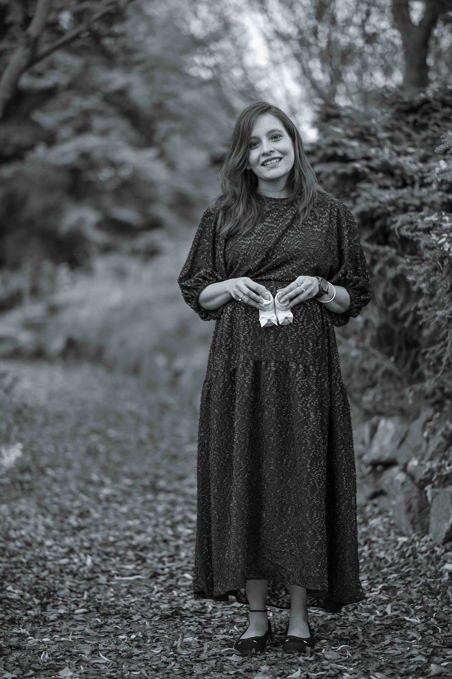 Smiling pregnant woman in a dark floral dress holding baby shoes on a forest path.