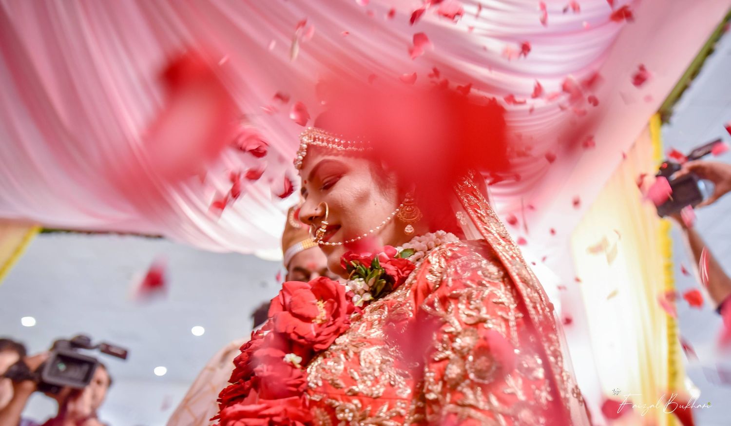 Indian bride in ornate red and gold lehenga surrounded by falling rose petals during wedding ceremony. Kamloops Wedding Photographer