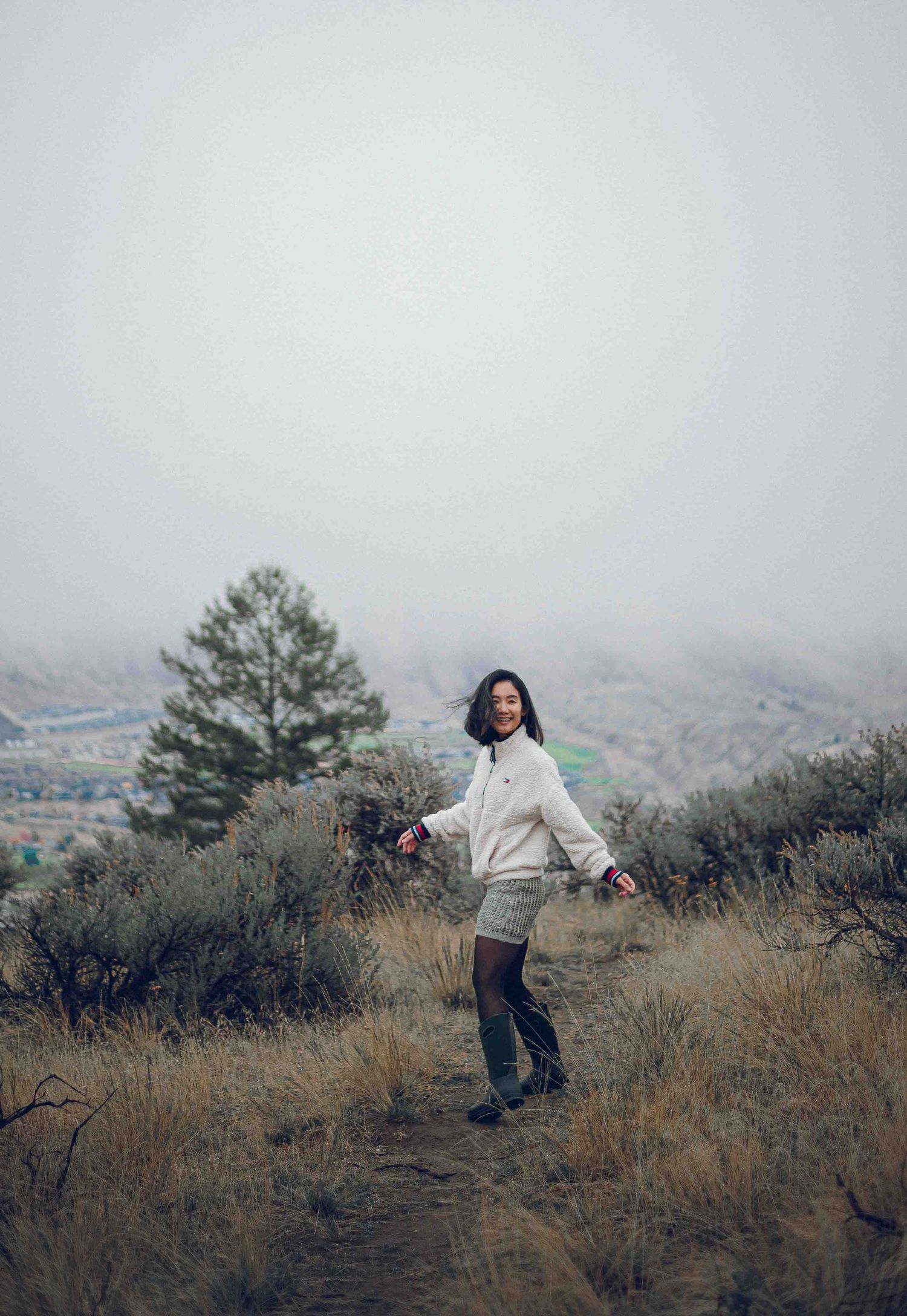 A smiling woman in a white sweater and shorts poses on a misty hiking trail surrounded by sagebrush.