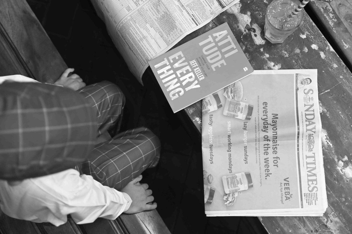 Close-up of a child in plaid pants sitting on a bench with Sunday Times and another newspaper.