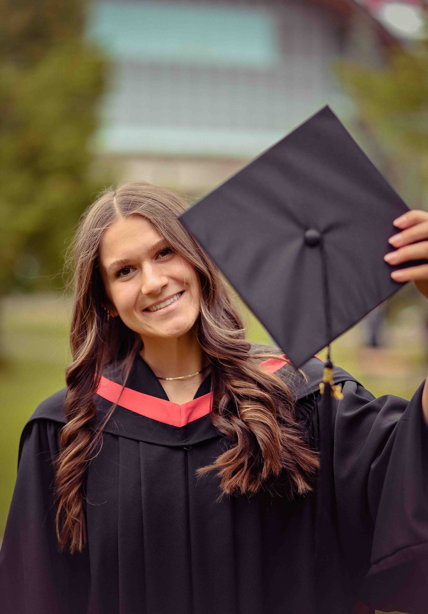 Smiling young woman in black graduation gown with red trim holding up her mortarboard cap outdoors. Best Graduation Photographer Kamloops
