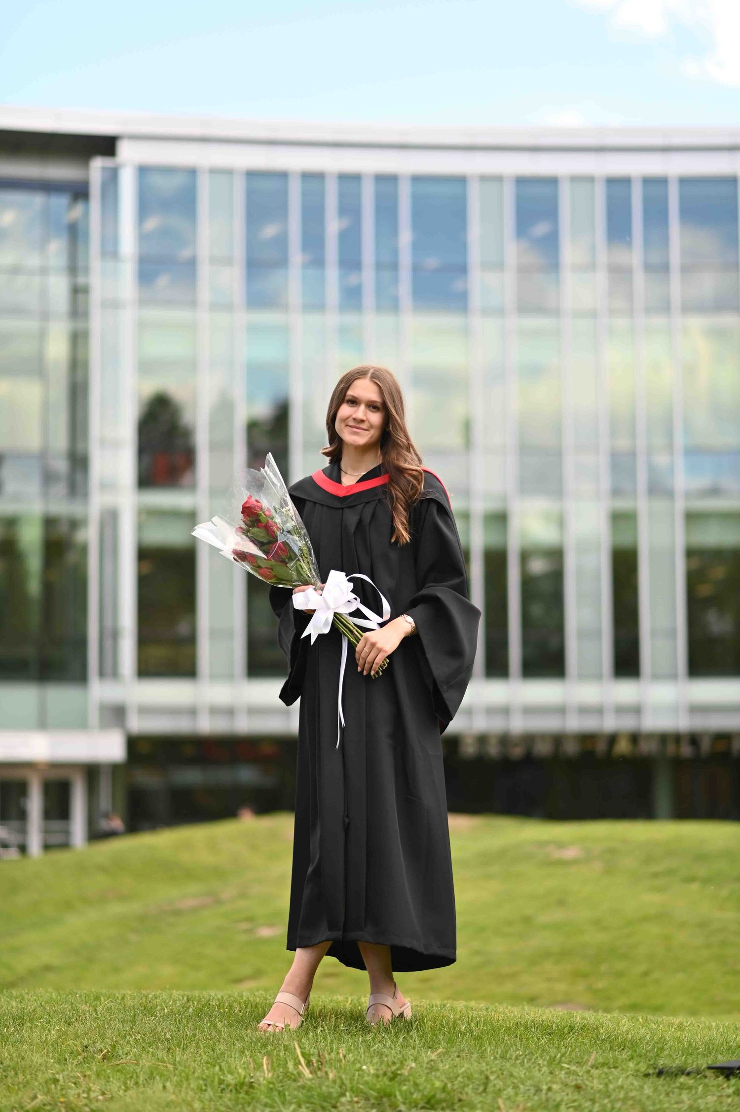 Young woman in black graduation gown holding bouquet of flowers standing on green grass outside university building.