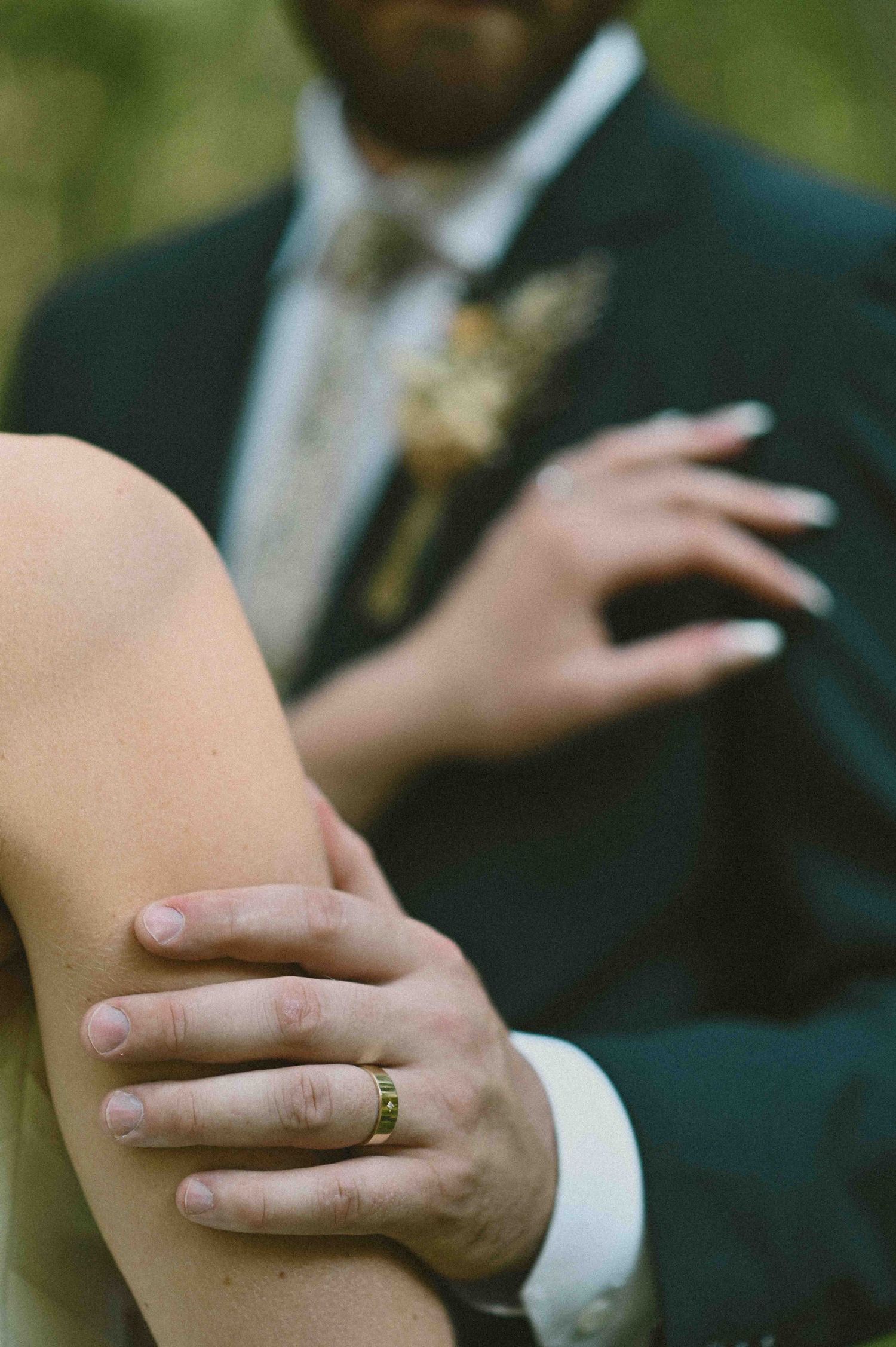 A groom in a navy suit gently holds a bride's arm, showcasing his gold wedding band.