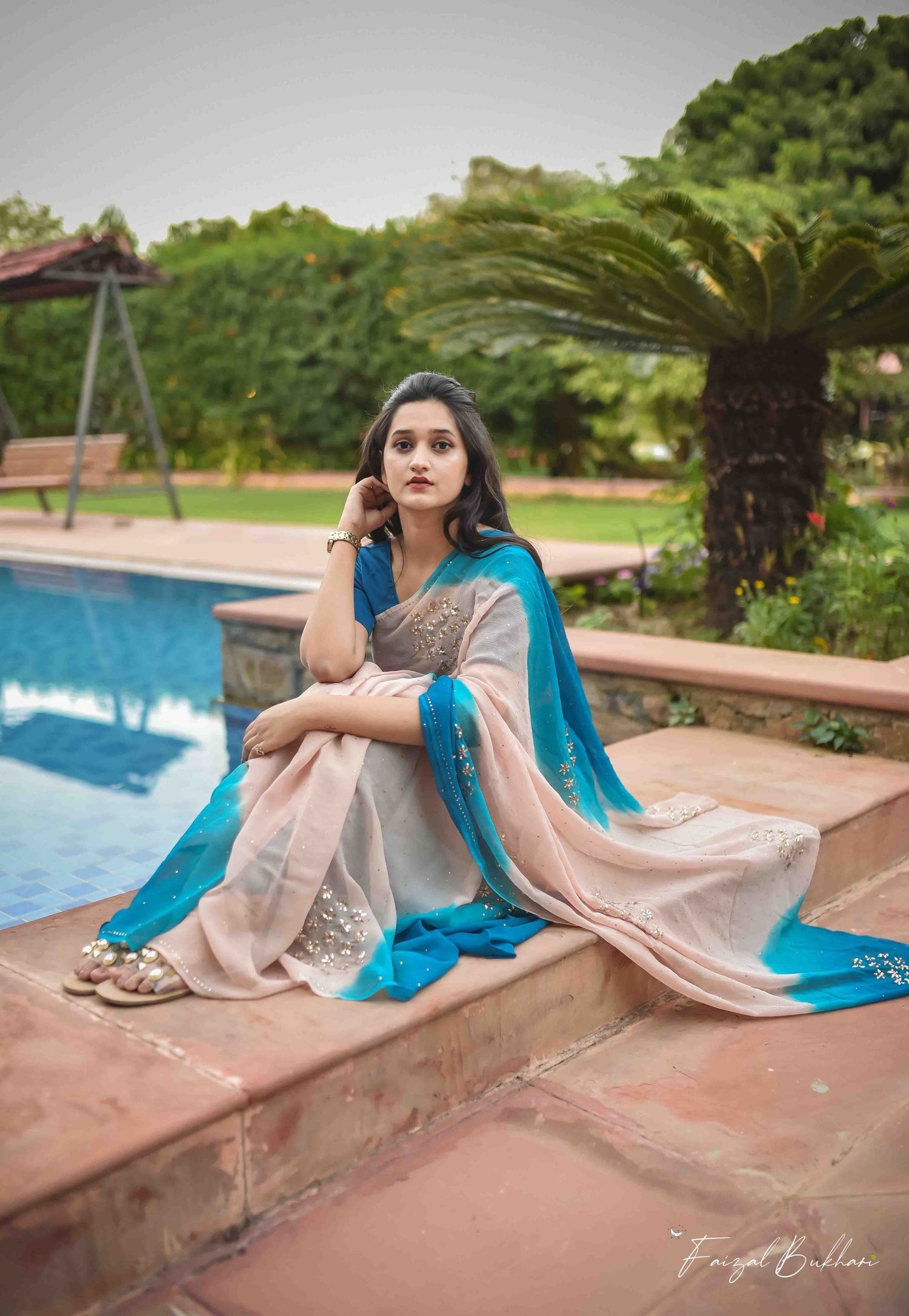 A woman in a gray and blue saree sits elegantly beside a pool in a lush garden setting.