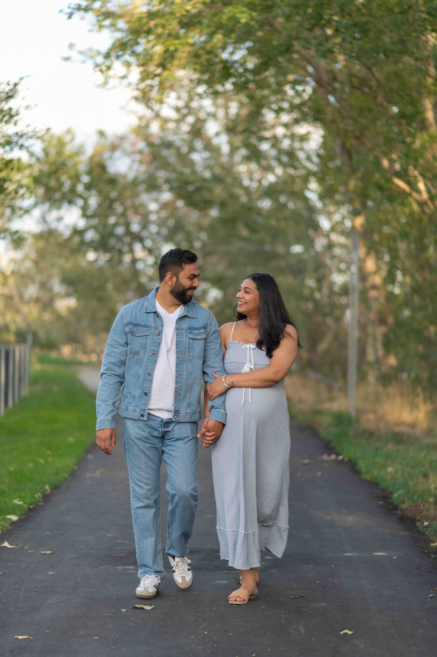 Expecting couple walking hand-in-hand on a scenic park path, woman in grey maternity dress smiling.