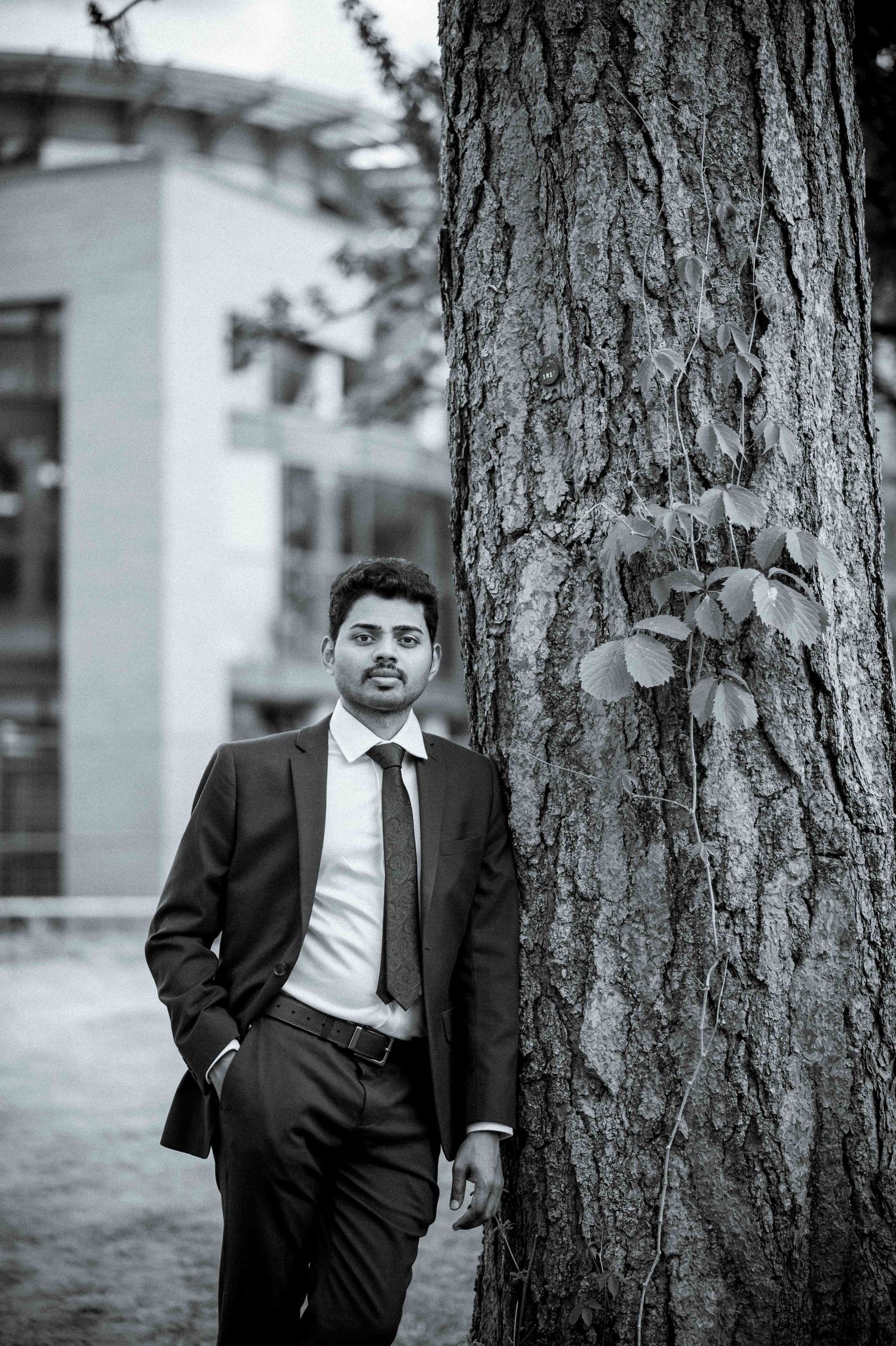 Black and white portrait of a man in a suit leaning against a tree on a campus.