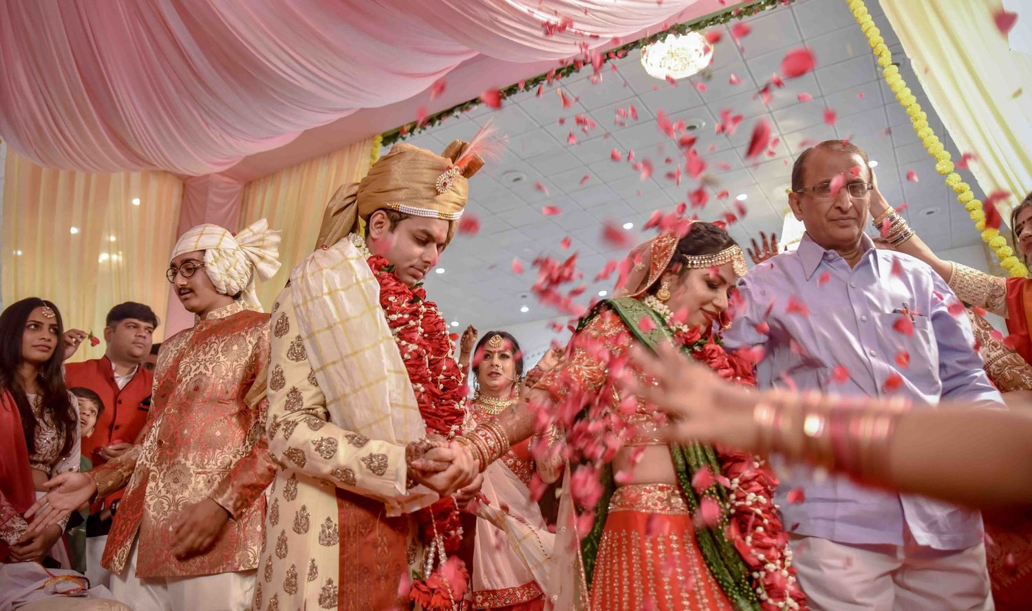 A vibrant Indian wedding ceremony with couple in red and gold attire amid flower petal shower.