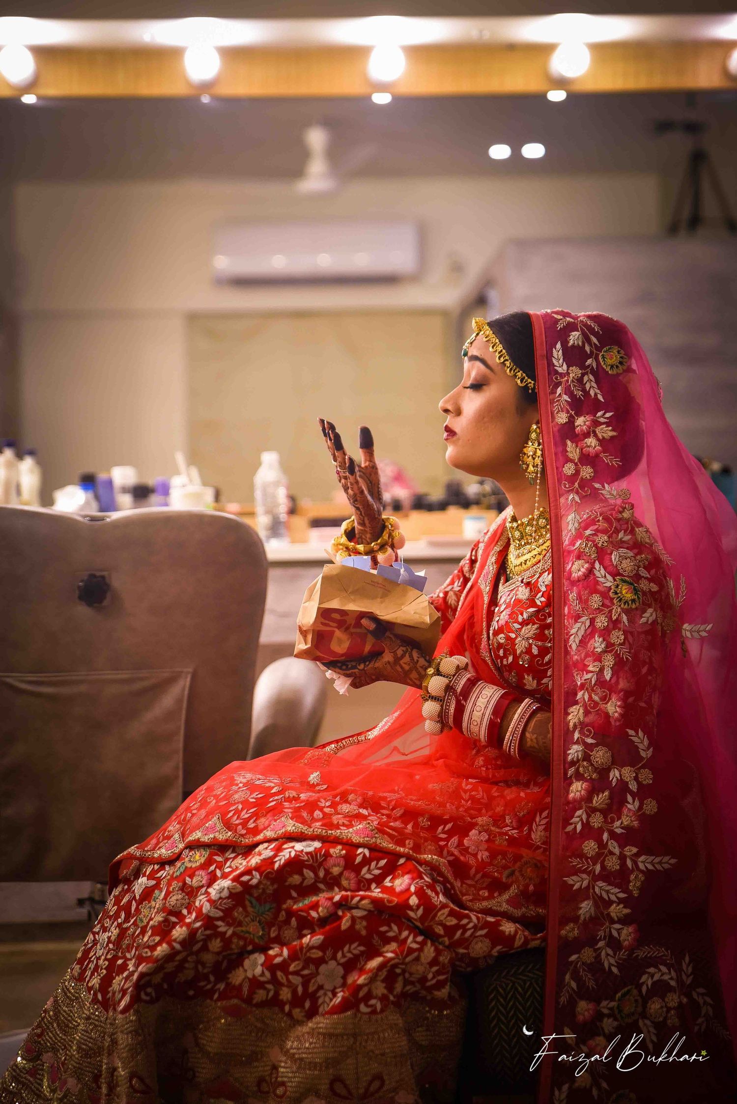 Indian bride in red bridal lehenga with gold jewelry and dupatta sitting elegantly during pre-wedding preparations.