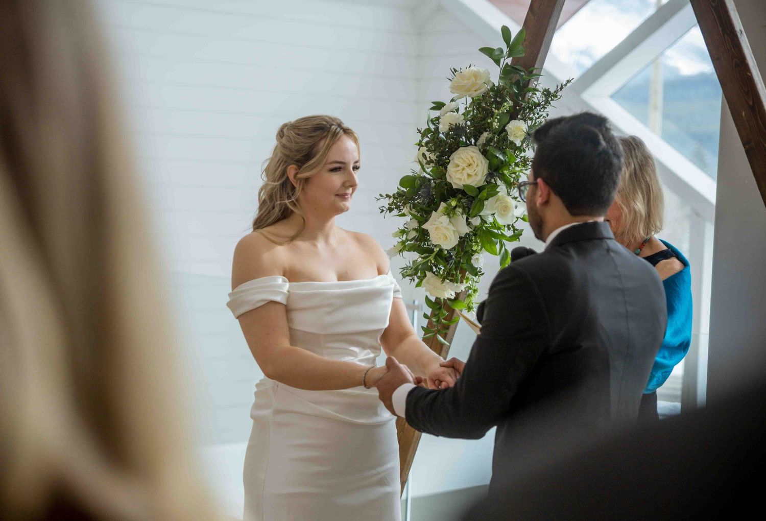 Bride in off-shoulder white gown holding hands with groom during indoor wedding ceremony beside floral arrangement.