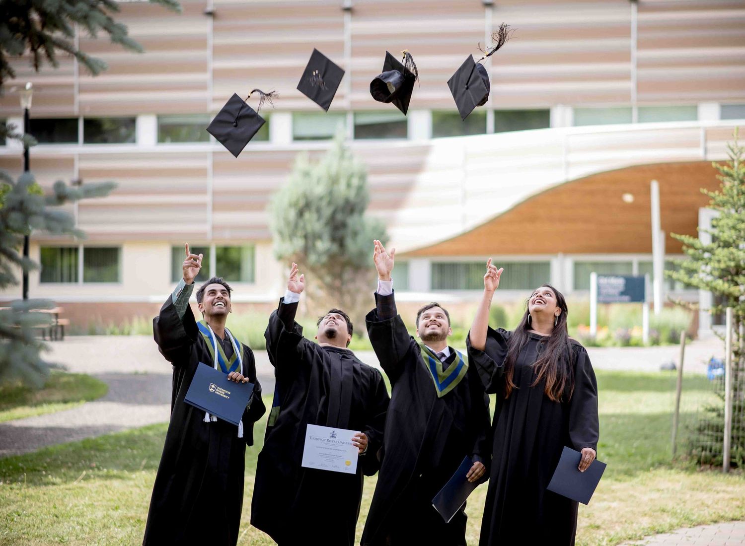 Four graduates in black robes toss their caps into the air outdoors, celebrating their graduation day.