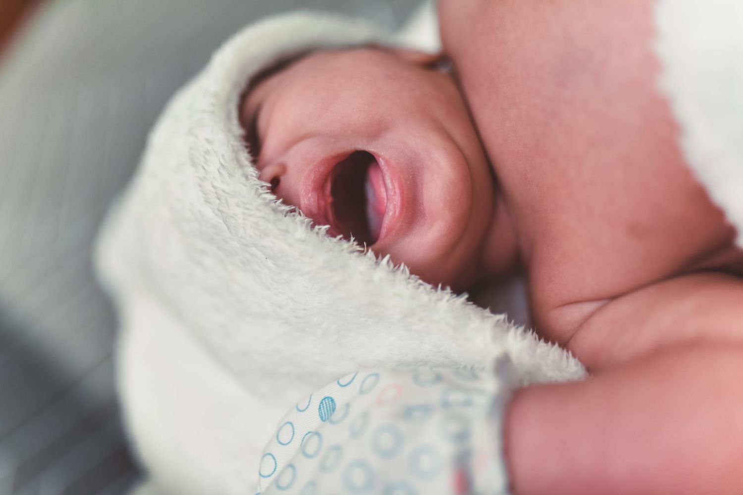 A newborn baby wrapped in a white hospital blanket with mouth open, captured in soft focus.