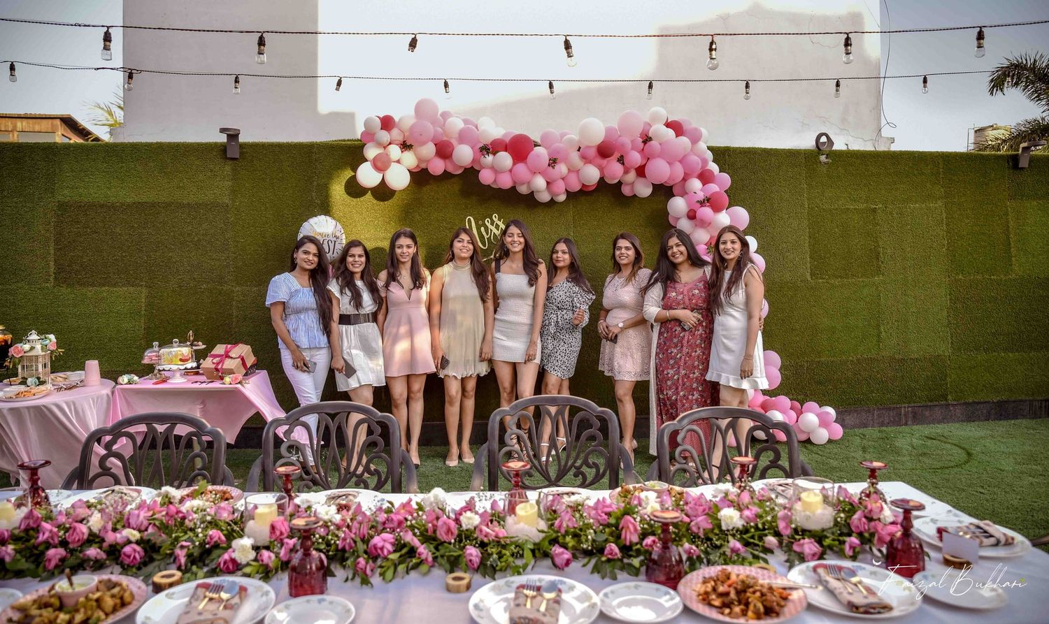 Bridal shower group photo with women in pink outfits posing behind a floral-decorated table and balloon arch.