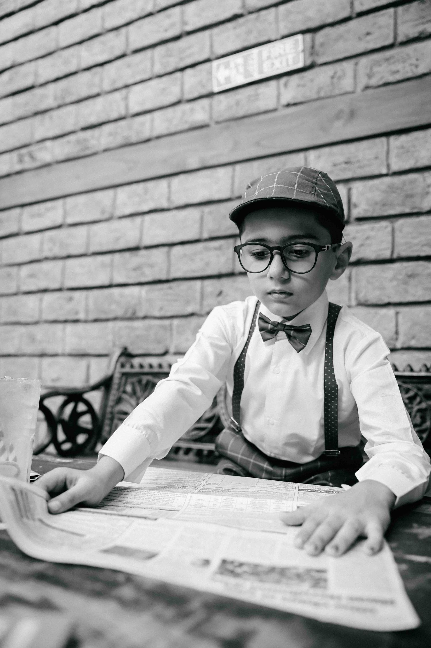 A stylish young boy wearing suspenders, bow tie, and cap reads a newspaper at a table by a brick wall.