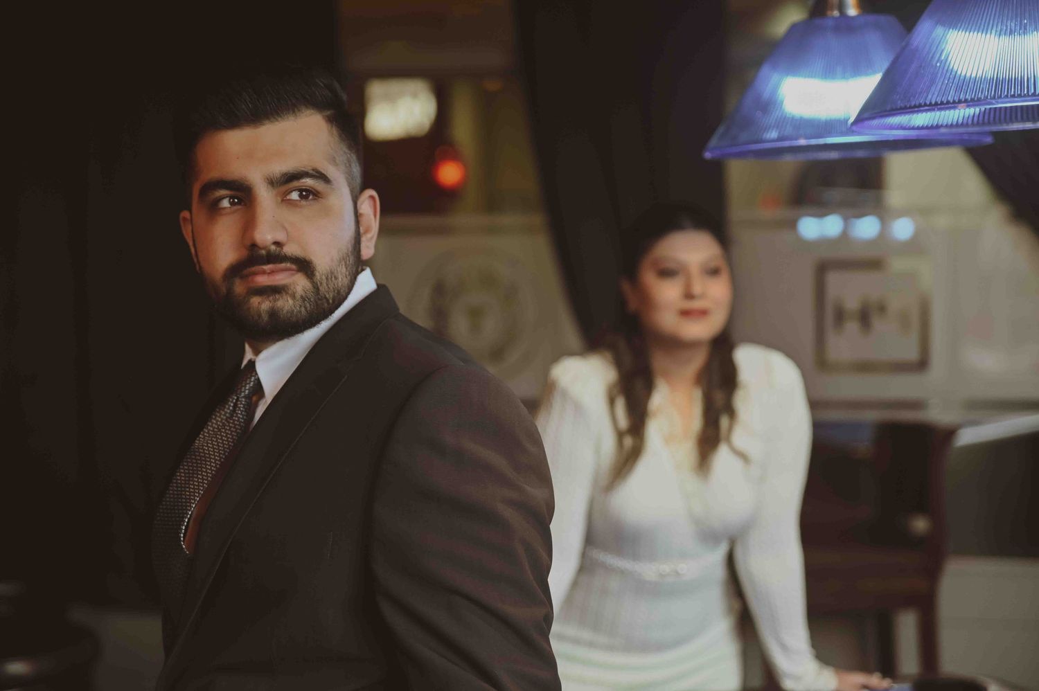 Stylish couple poses in a dimly lit restaurant, man in suit and woman in white dress for pre-wedding shoot.