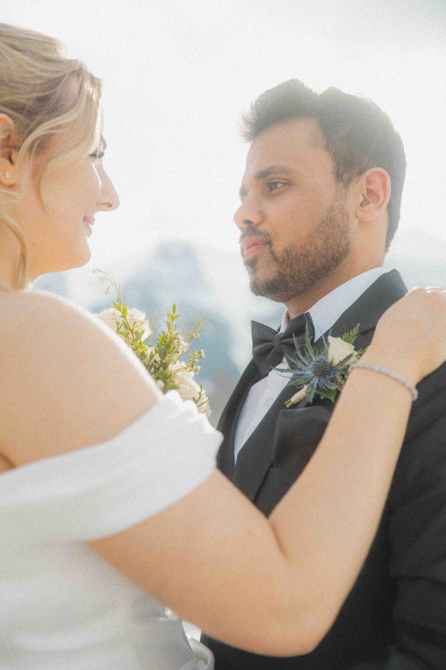 Bride and groom share intimate moment, she in off-shoulder white dress holding bouquet, he in black tuxedo with boutonniere.