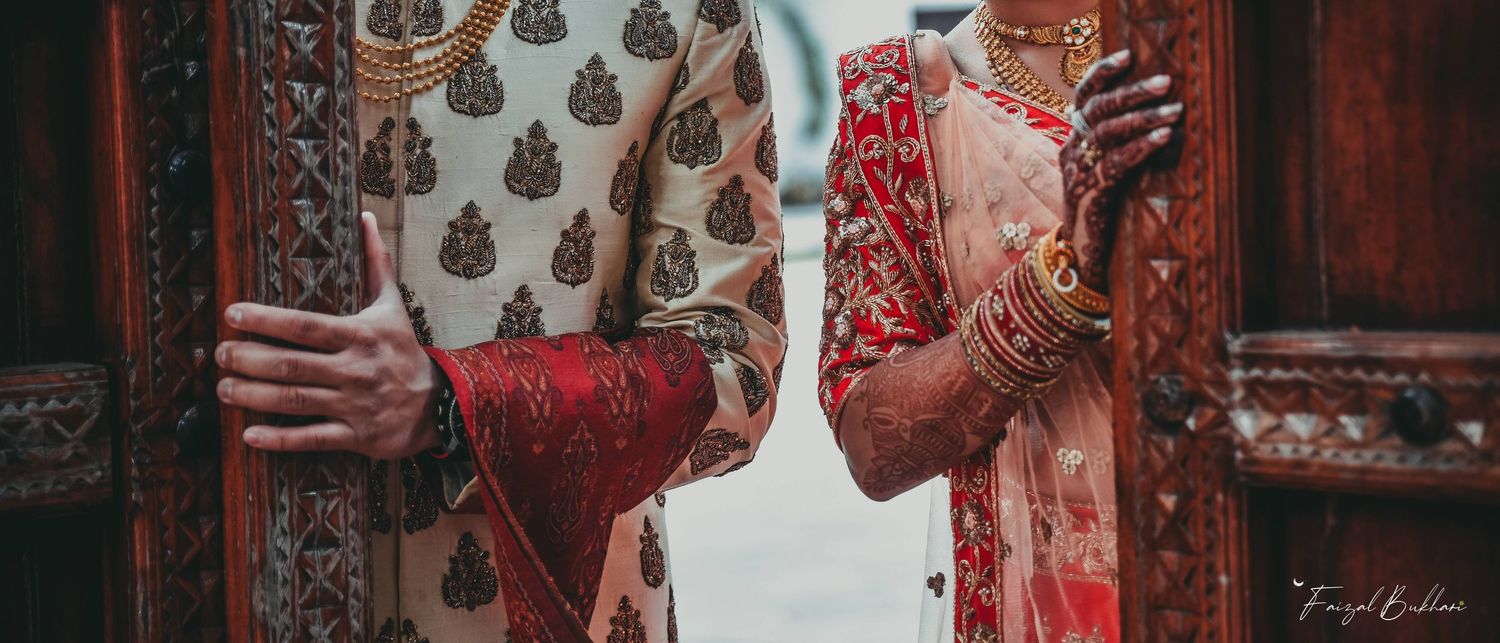 Indian bride and groom in traditional red and gold wedding attire standing together at ornate wooden doors.