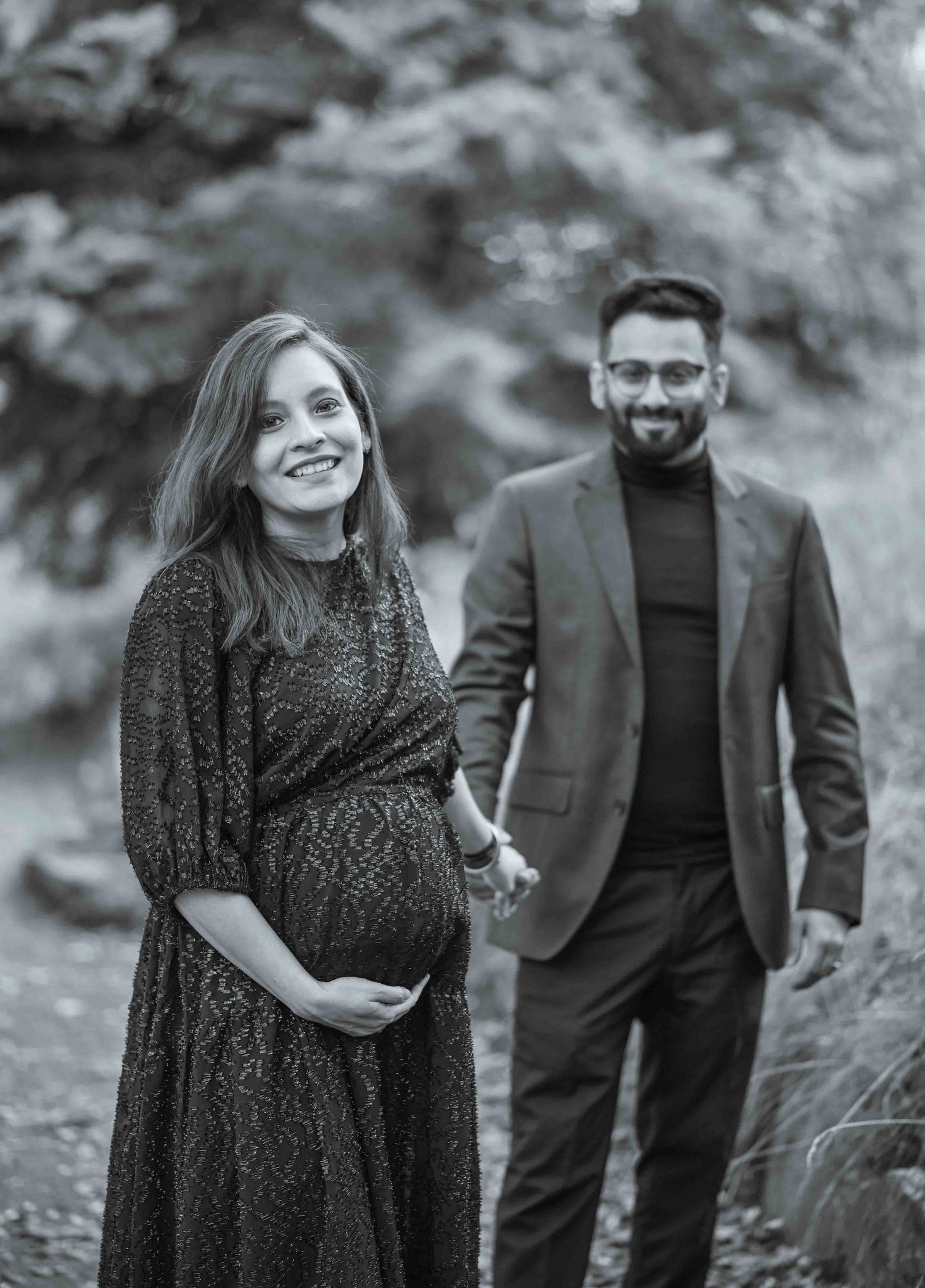 Black and white maternity photo of a couple holding hands on a scenic outdoor path.