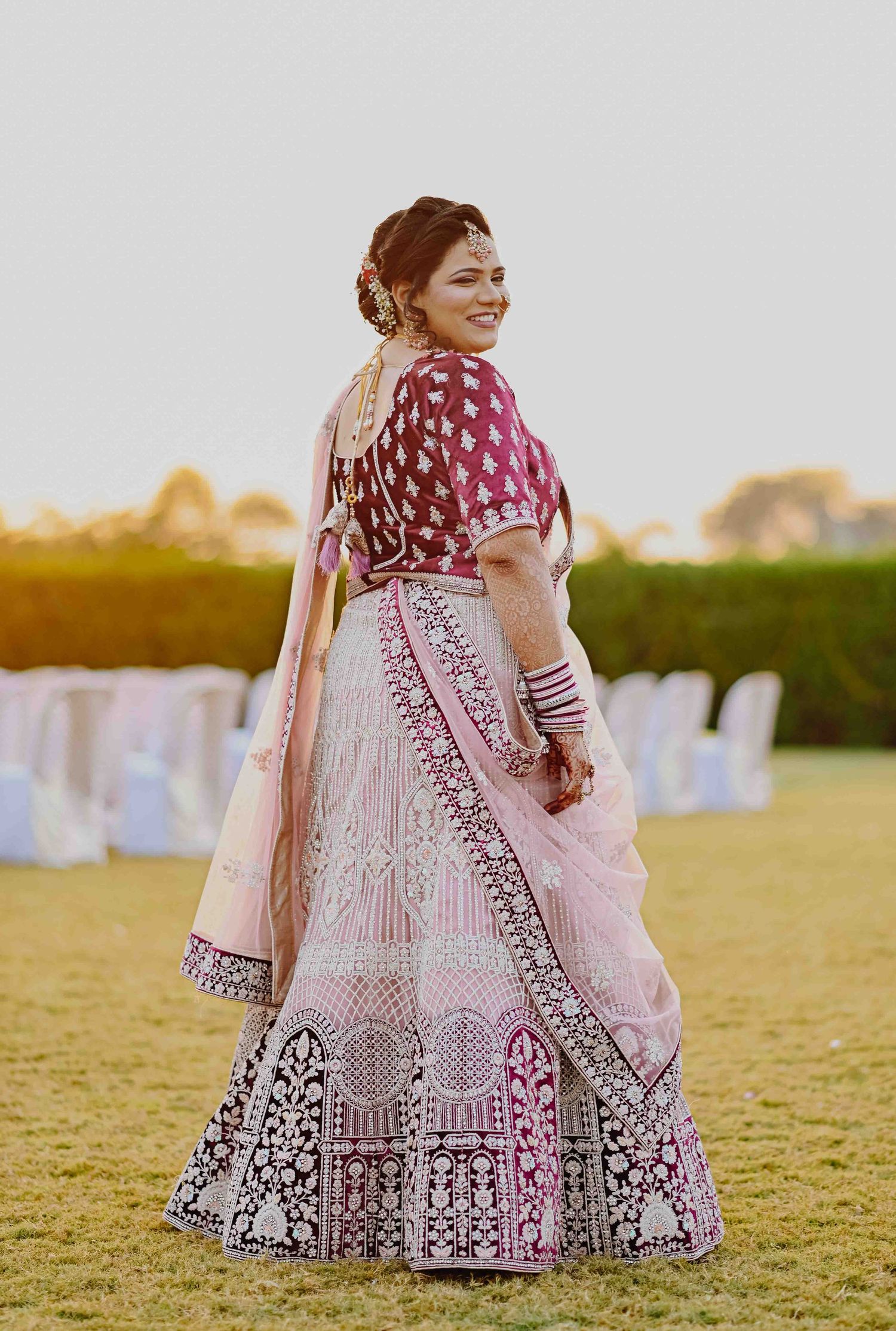 Indian bride in pink and magenta lehenga smiling over shoulder at outdoor wedding venue.
