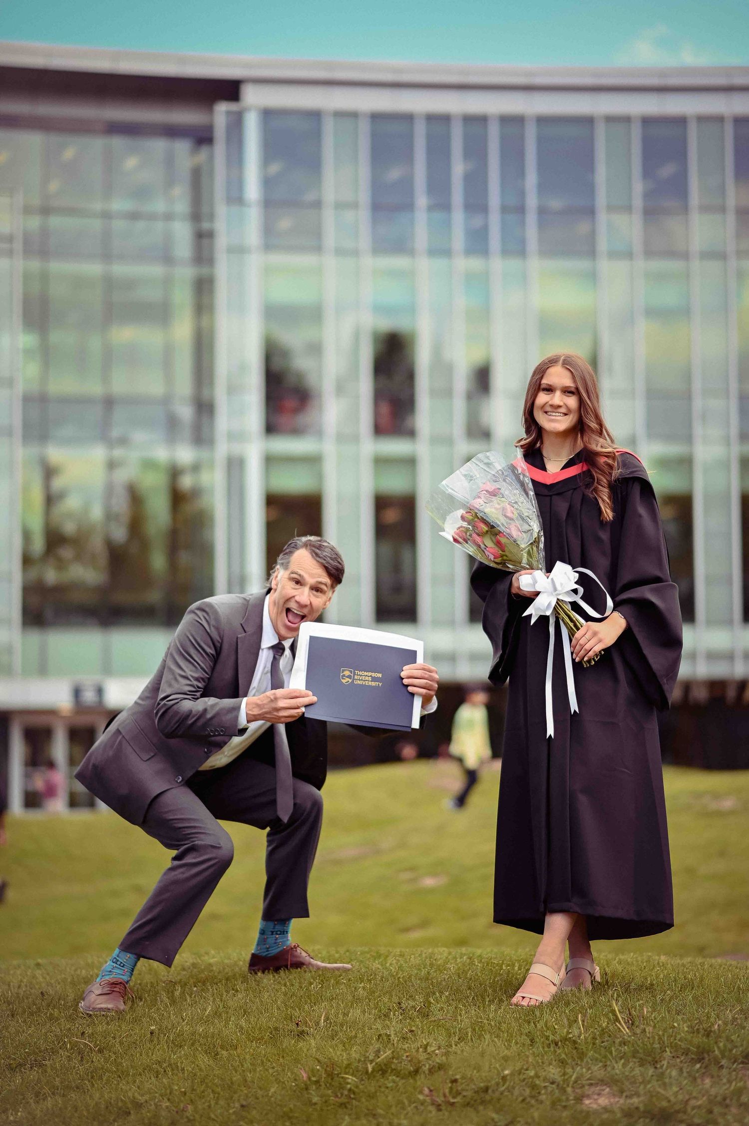 Proud father humorously shows TRU diploma while daughter in graduation gown holds flowers outside.