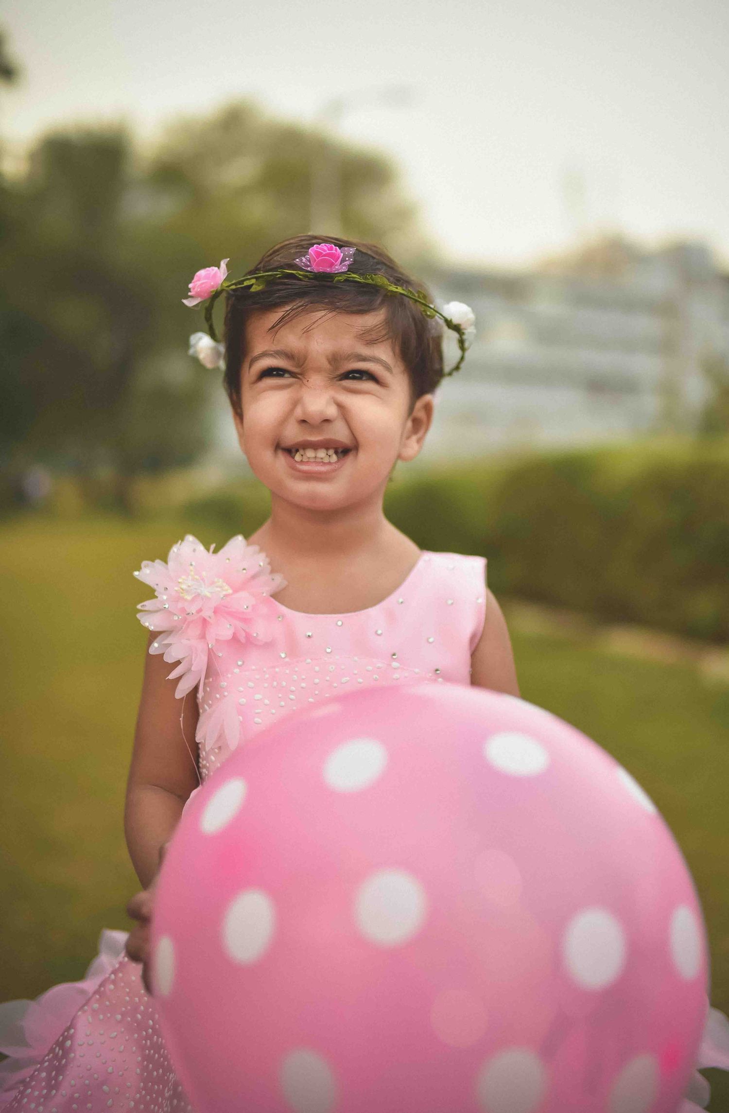 Smiling little girl in pink dress and floral crown holding a pink polka dot balloon outdoors.