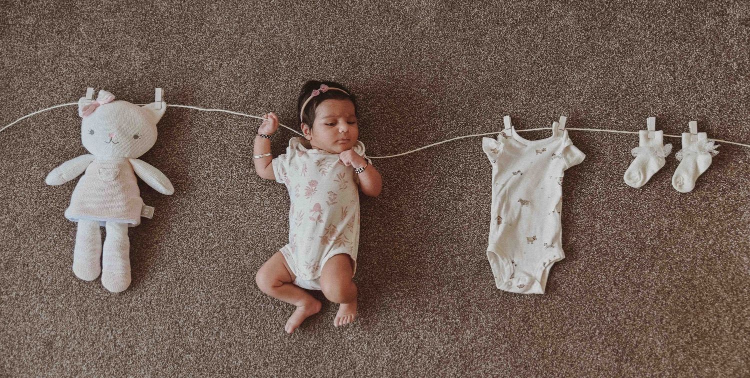 Newborn baby in white onesie lying on floor beside stuffed bunny and mini clothes on a decorative line.