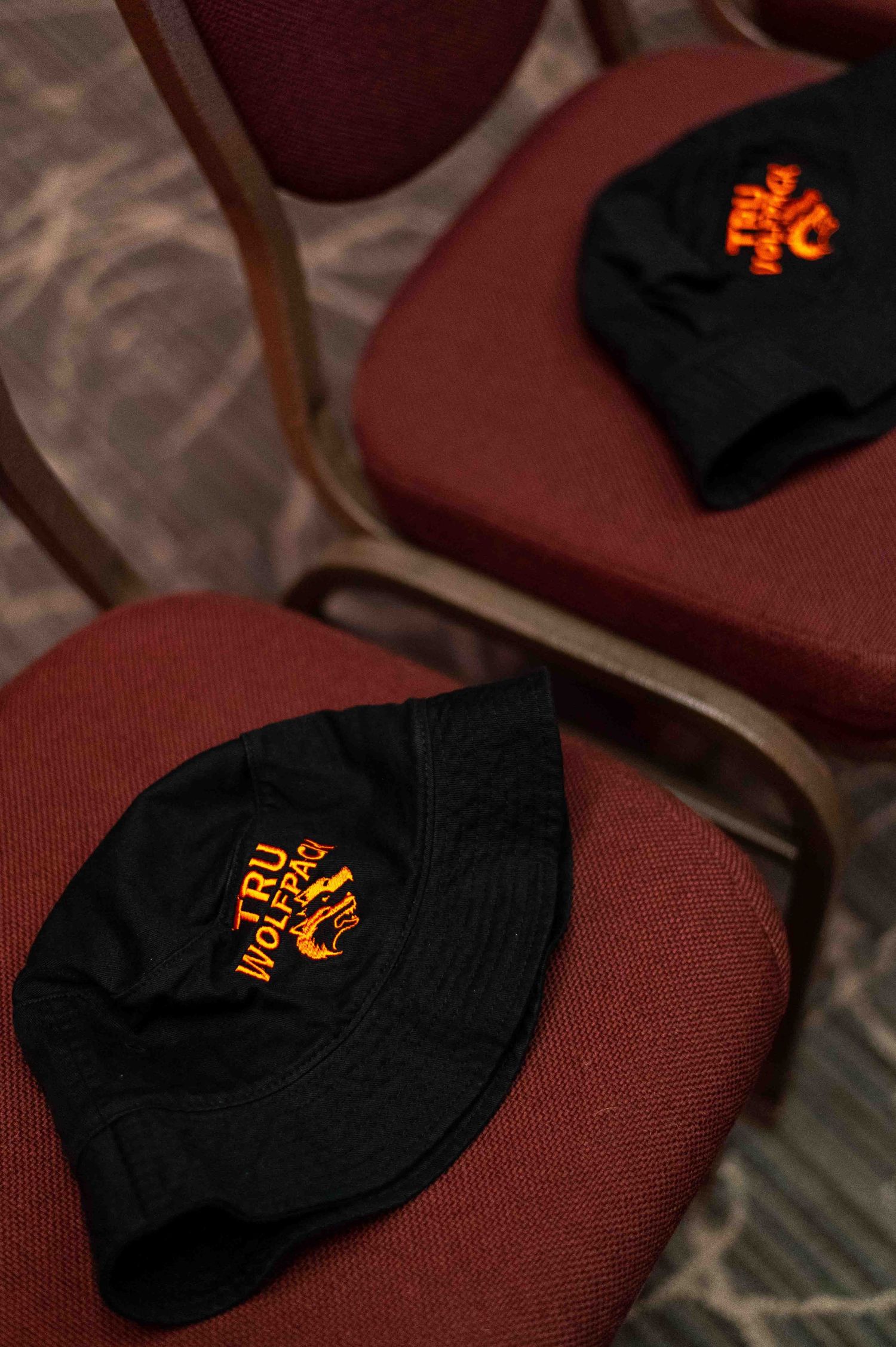 Two black TRU WolfPack embroidered bucket hats resting on red cushioned chairs at an event.