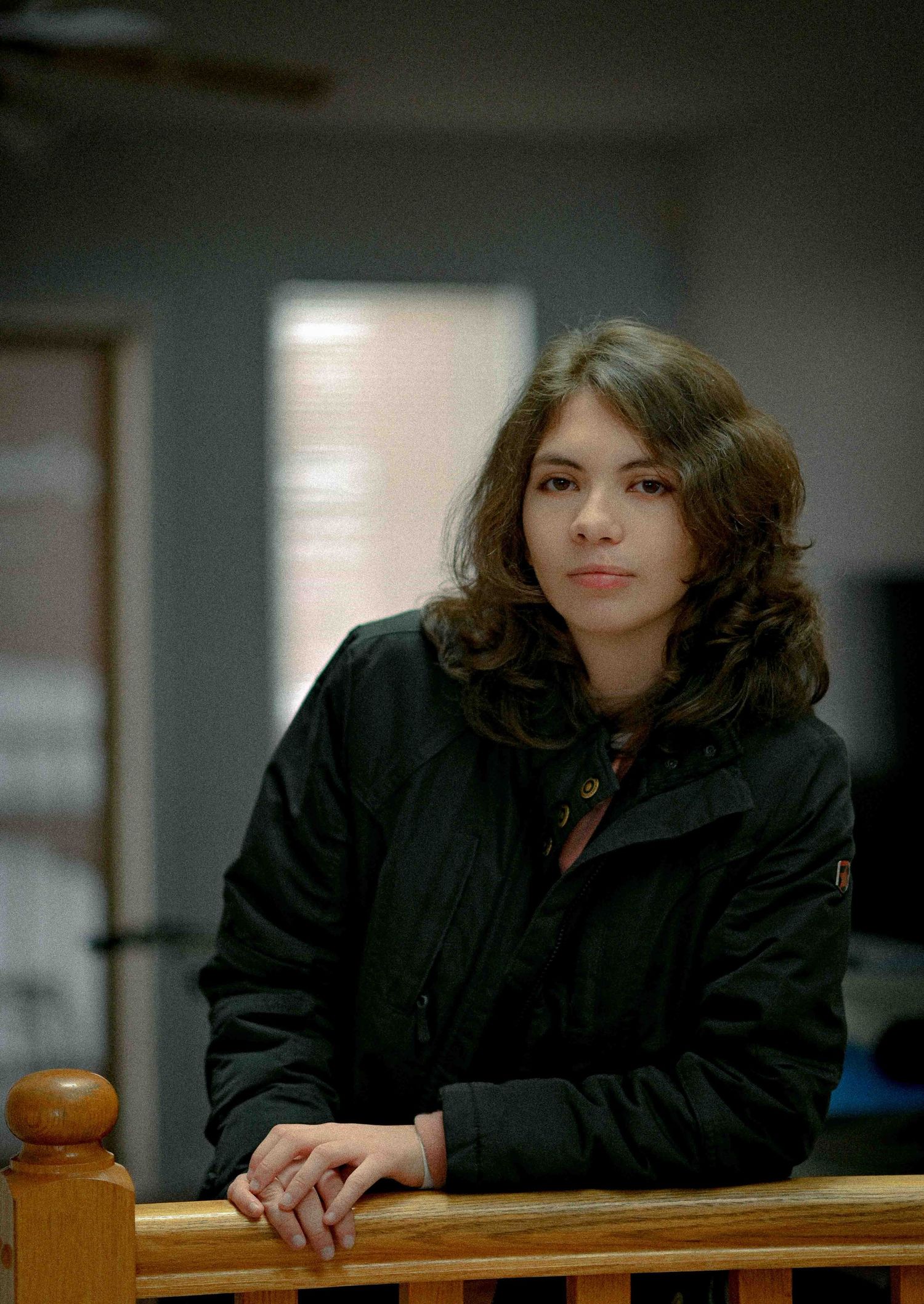 A young woman with wavy brown hair in a black jacket leans on a wooden railing indoors.