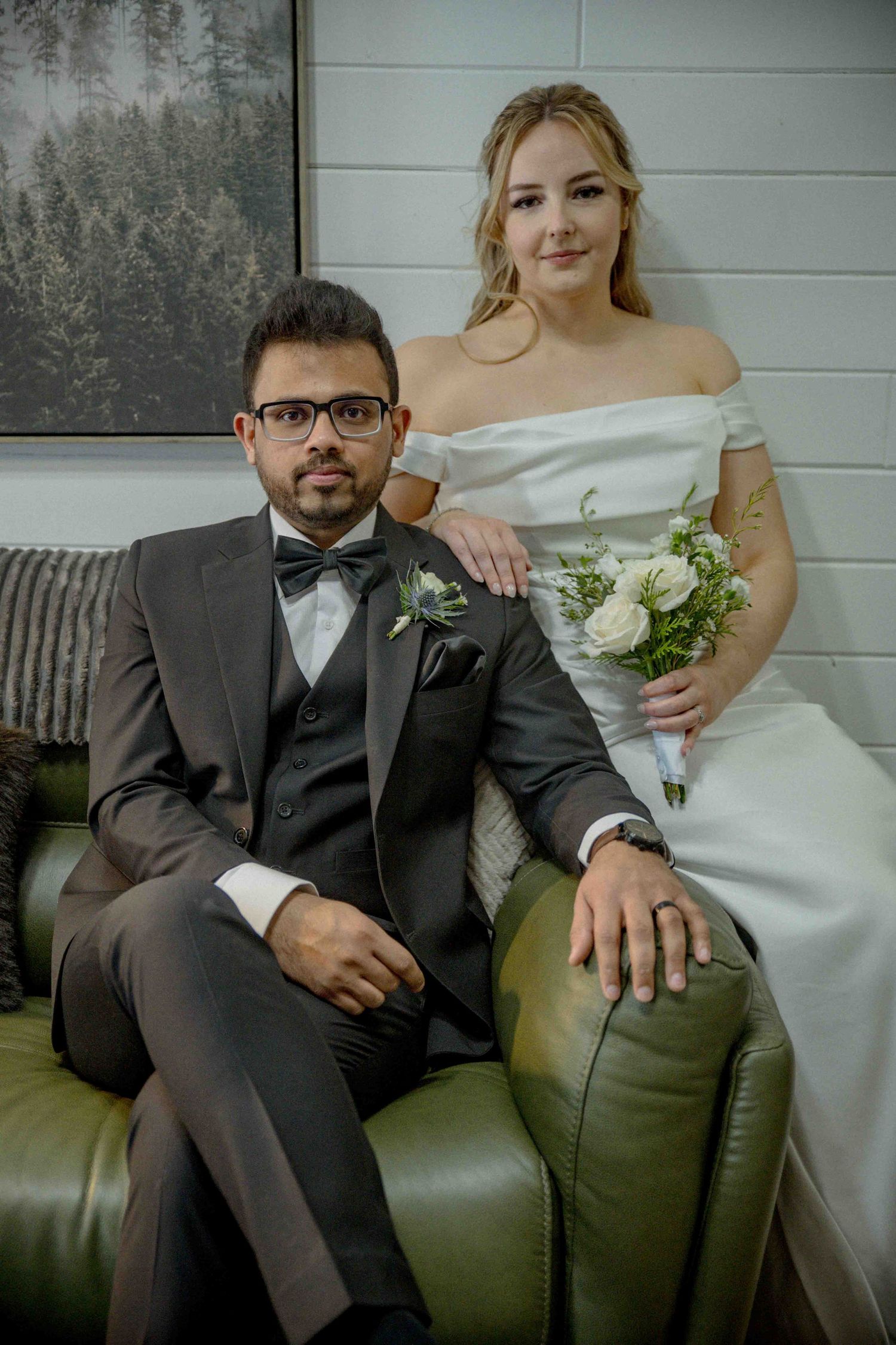Bride in off-shoulder white gown with bouquet poses with groom in gray three-piece suit on green leather sofa.