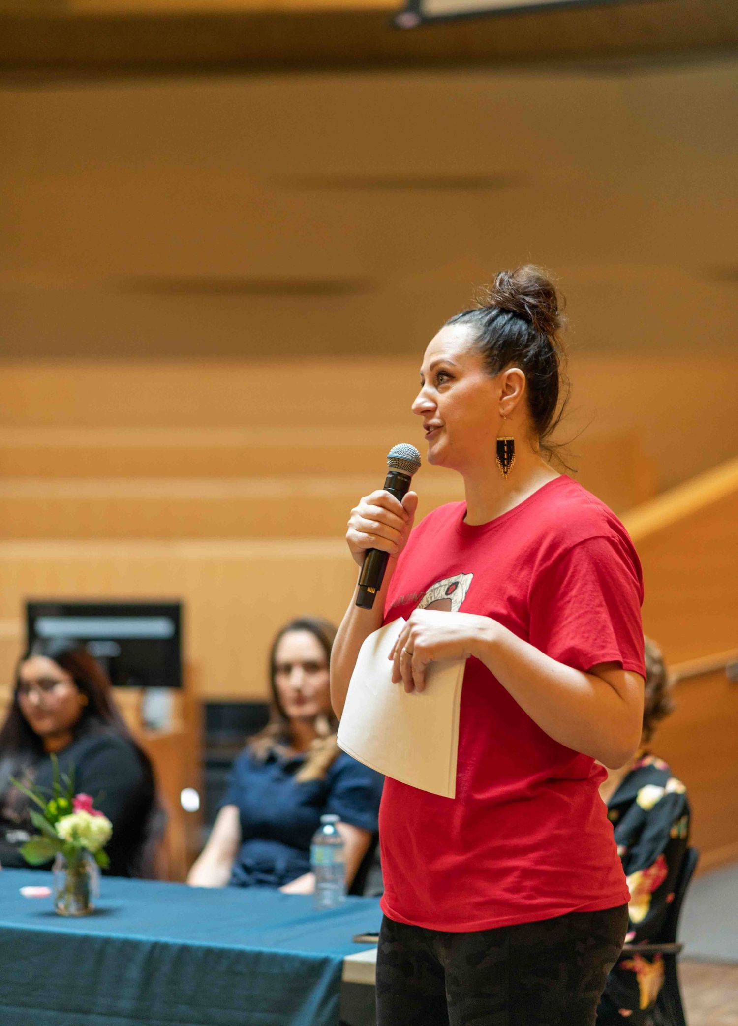 Woman in red shirt speaks into microphone at indoor event while holding papers.