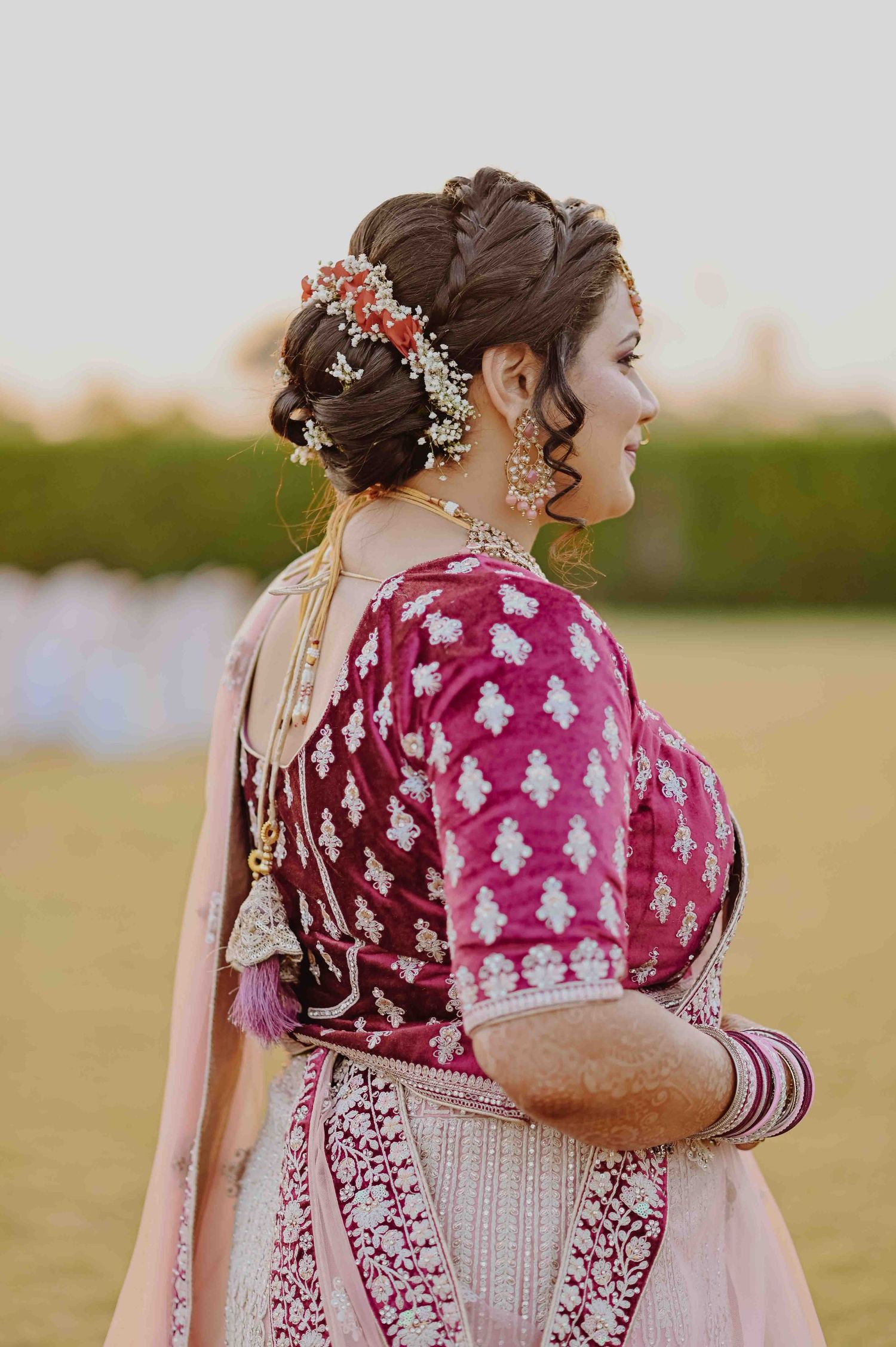 Indian bride in magenta embroidered blouse with floral bun, smiling over her shoulder.