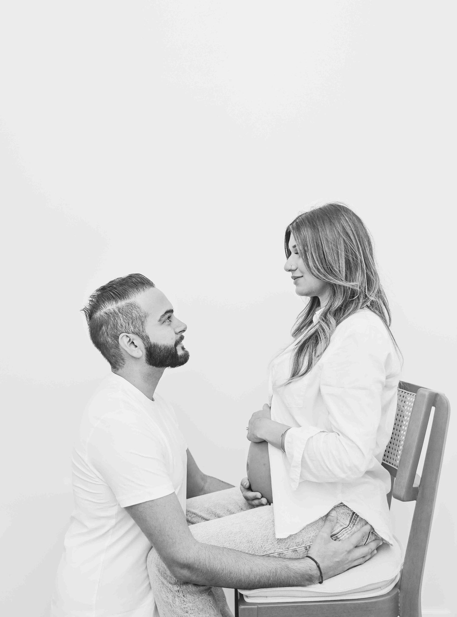 Black and white maternity photo of couple, man kneeling before seated pregnant woman in white shirt.