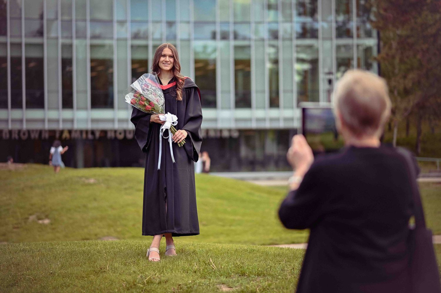 Graduate in black gown poses with white flowers outside Brown Family House of Learning building.