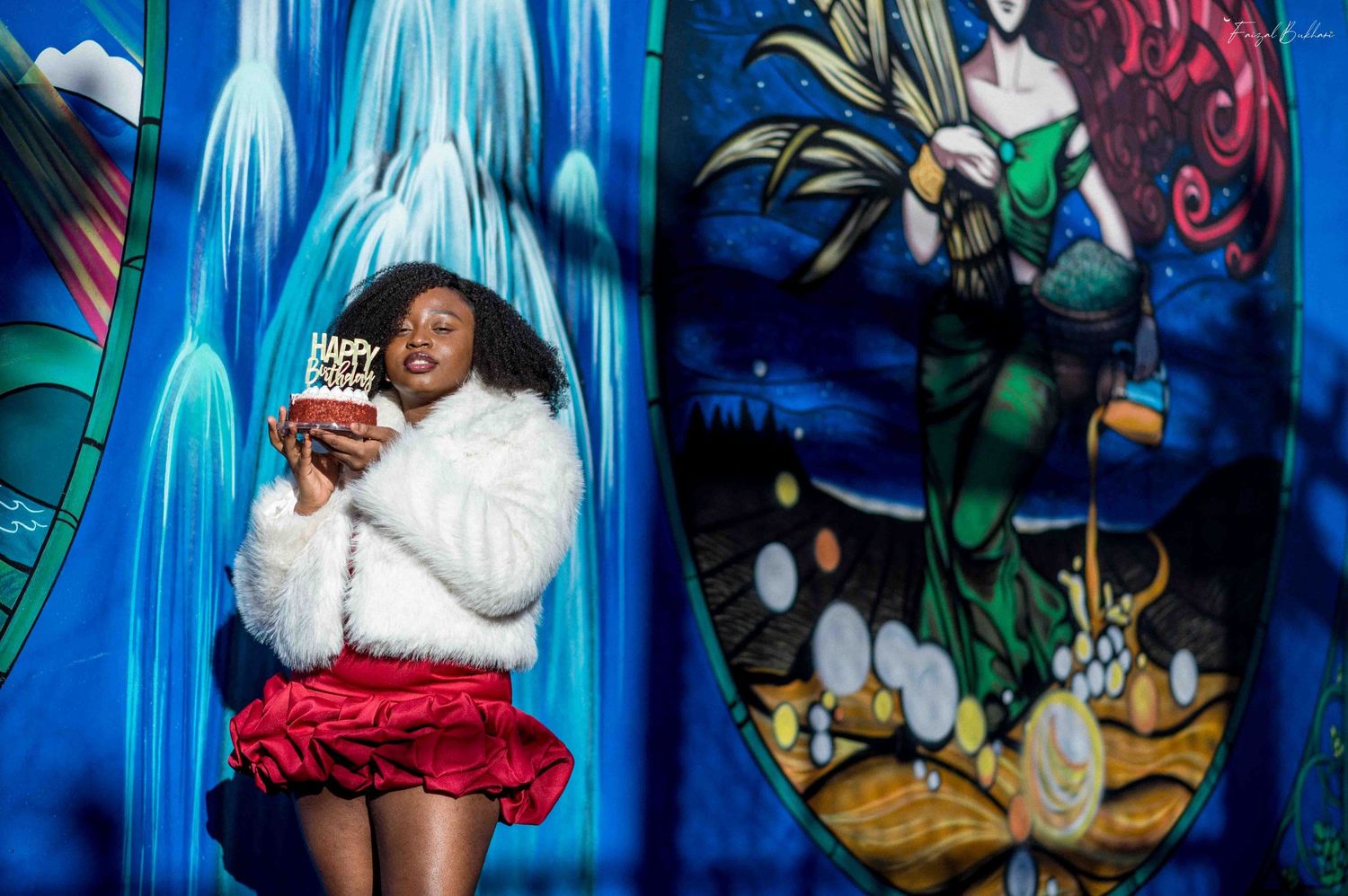 Woman in white fur jacket and red skirt holding birthday cake in front of vibrant mural wall.