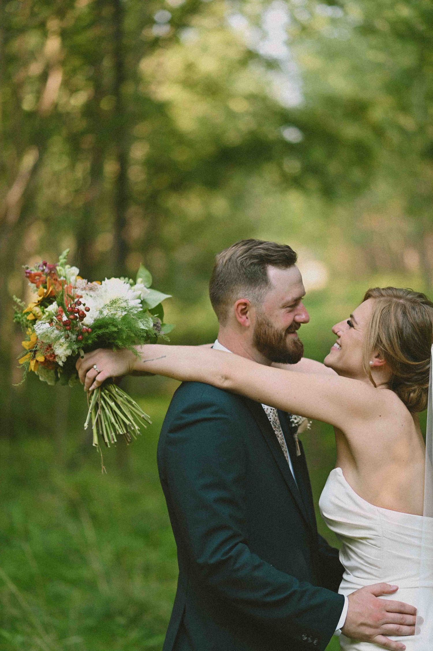 Bride hugs groom from behind holding colorful bouquet, both smiling joyfully in a lush green forest setting.