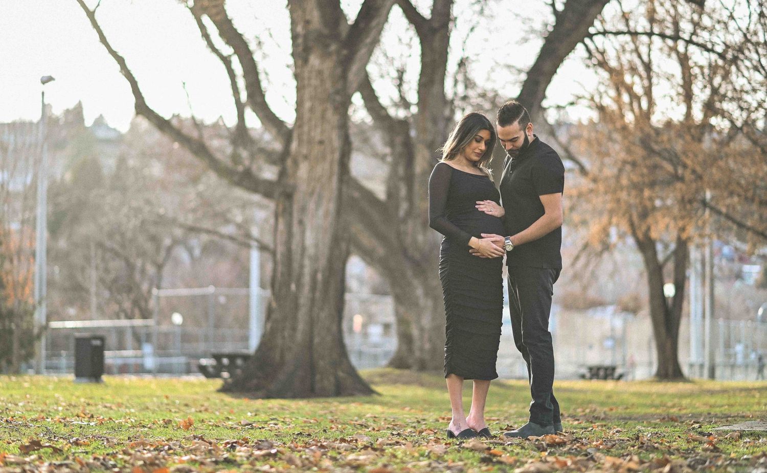 Couple gazing lovingly at pregnant belly under a tree in sunny park.